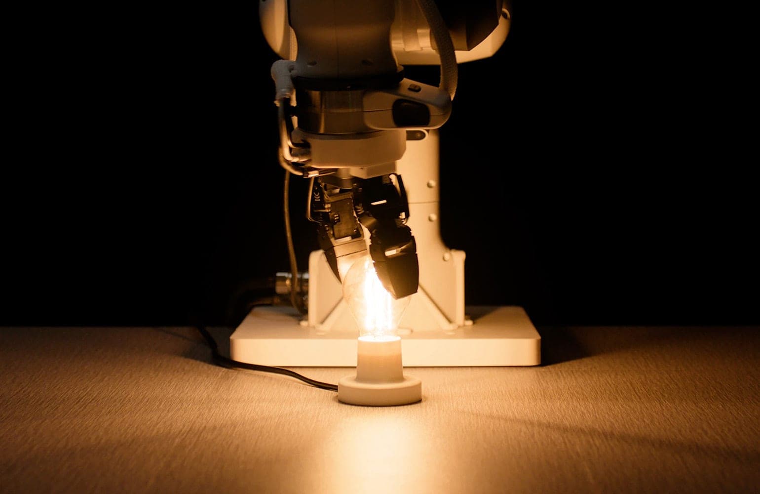 A robot arm with a two-finger gripper holding a glowing light bulb on a dark table
