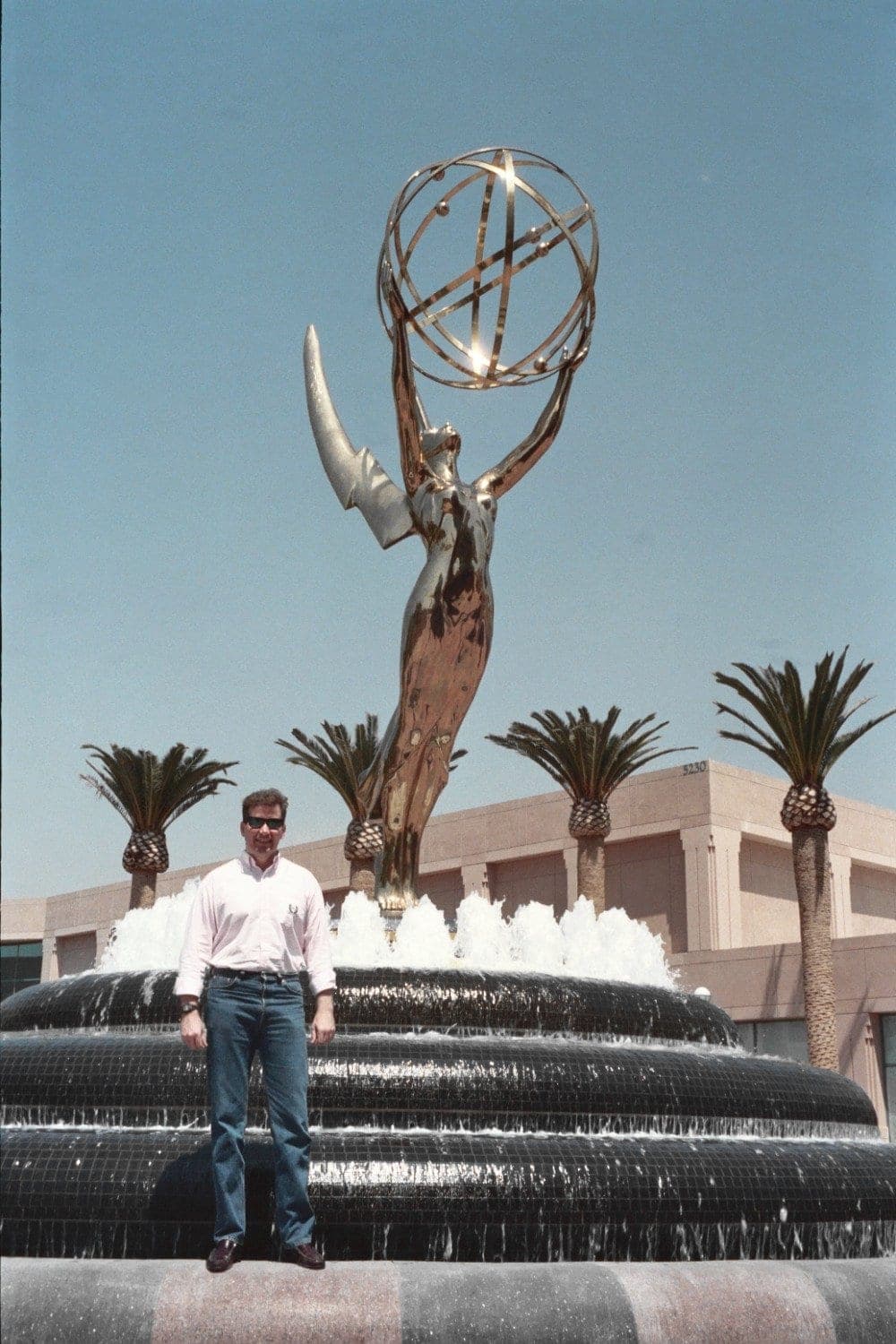 A man stands in front of a giant gold Emmy statue rising from a fountain, with palm trees and a building behind it