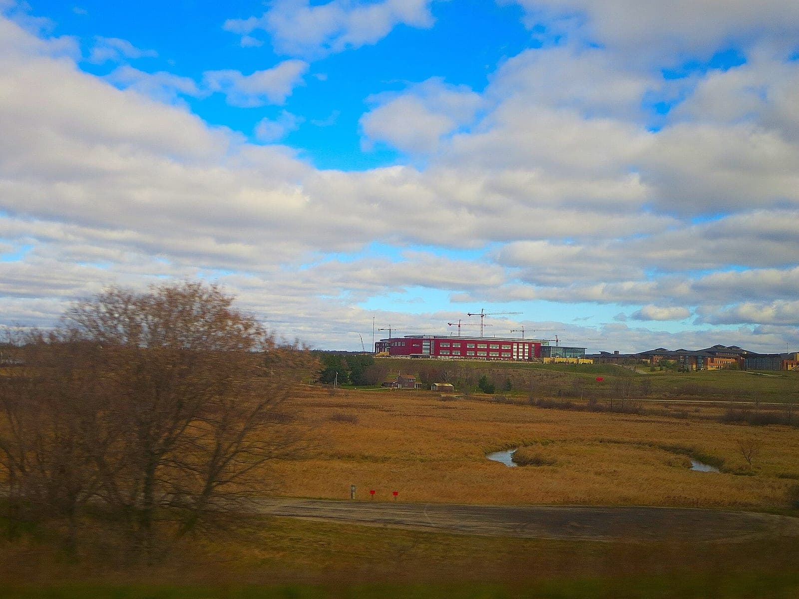 Epic Systems' campus buildings in Verona, Wisconsin, seen across a field and wetland under a cloudy blue sky, with construction cranes behind