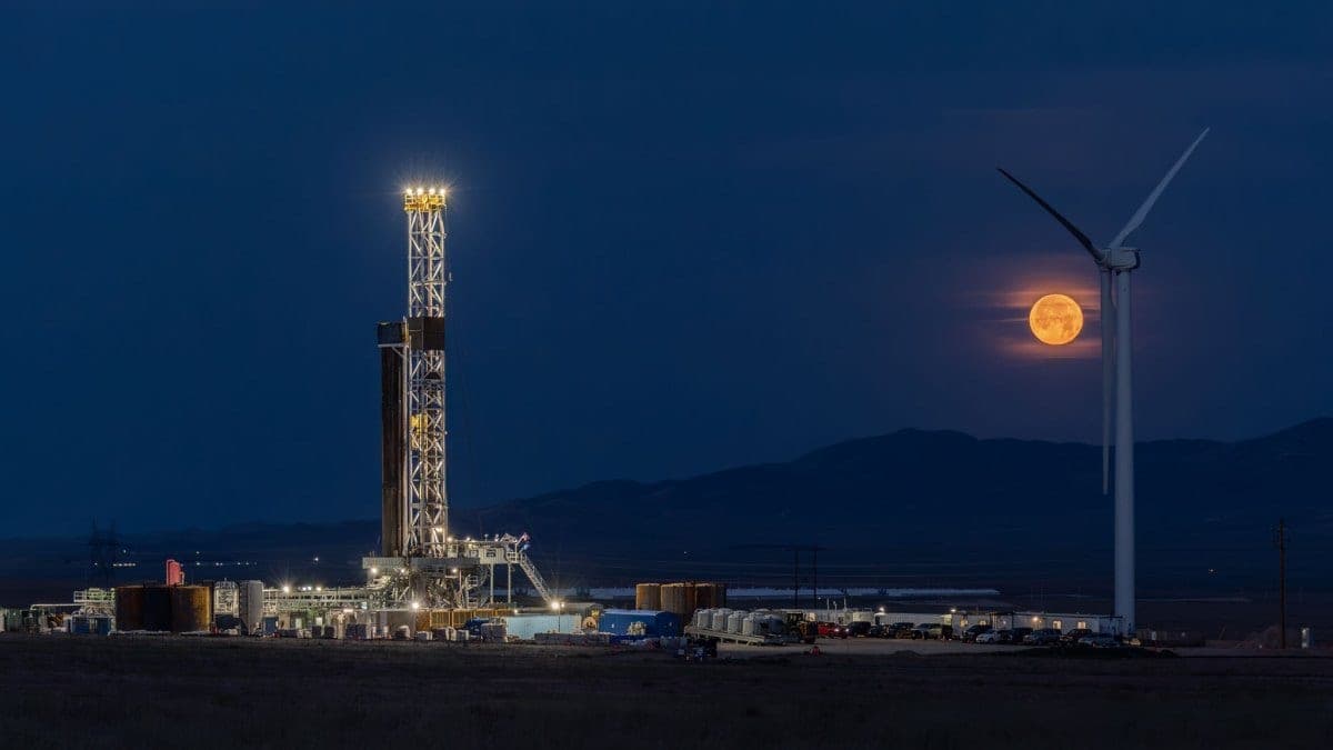 A brightly lit drilling rig at a Fervo Energy geothermal site at night, with a wind turbine and a full moon in the background
