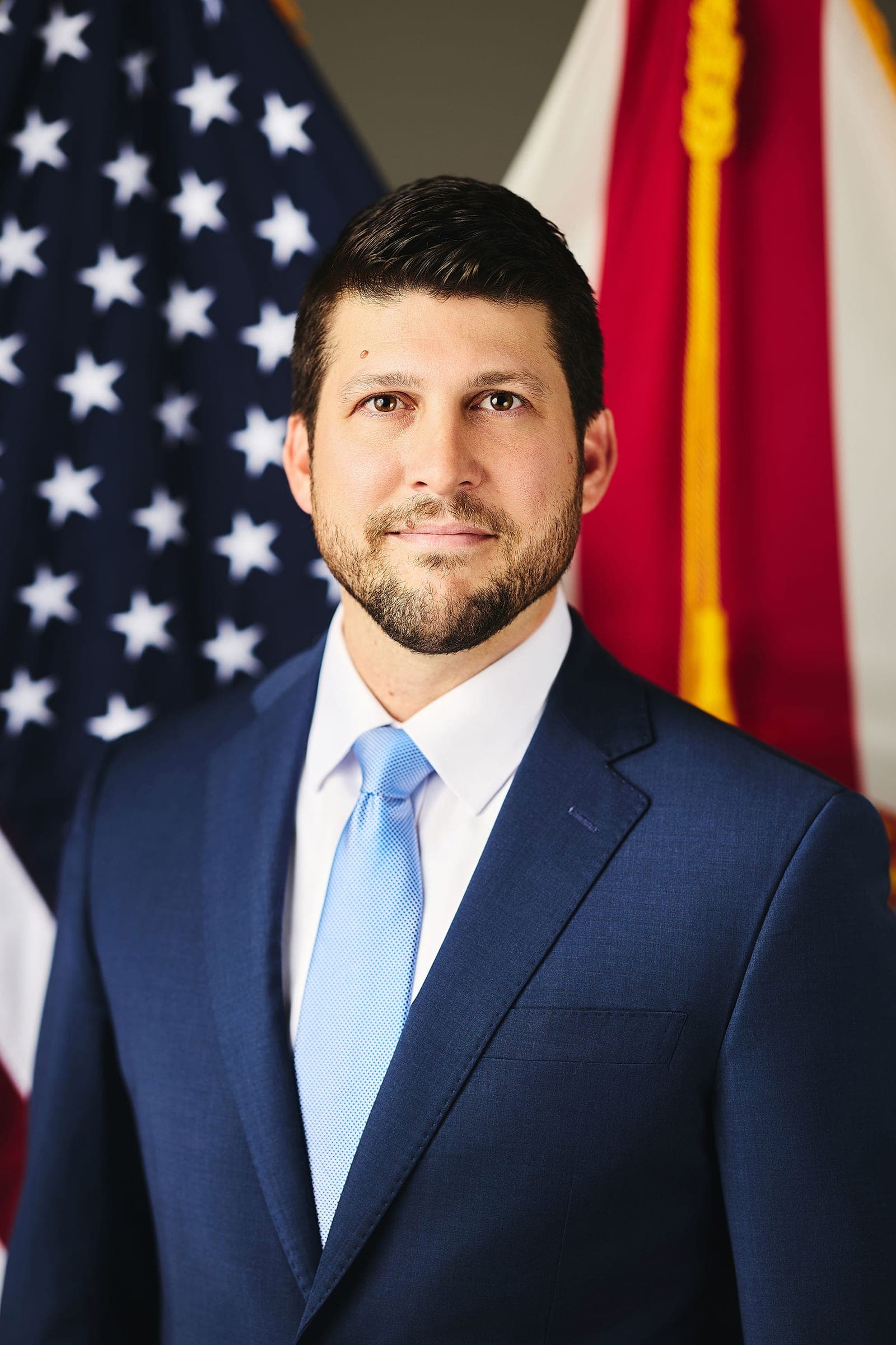 Official portrait of Florida Attorney General James Uthmeier in a navy suit and light blue tie, standing in front of American and Florida state flags.