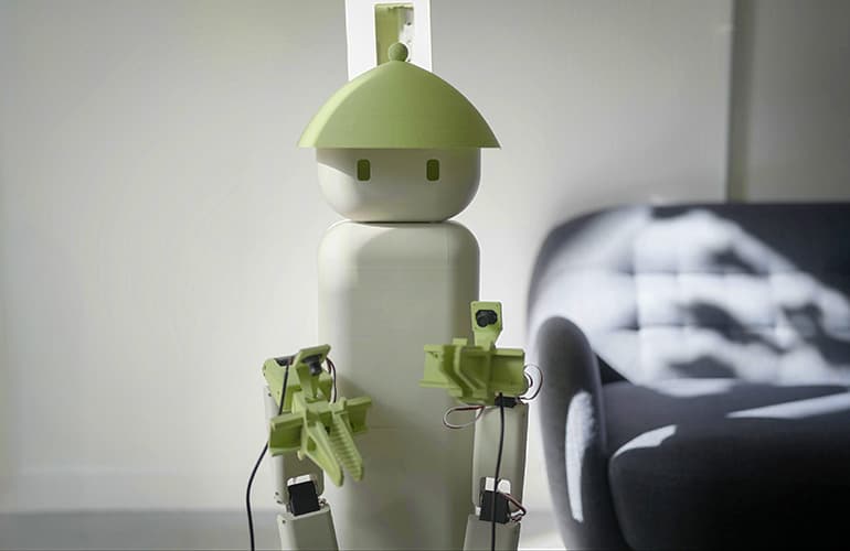 The Flourish One home robot, a pale green and white humanoid-style robot with a rounded head and two mechanical arms with grippers, standing in a living room next to a dark couch.