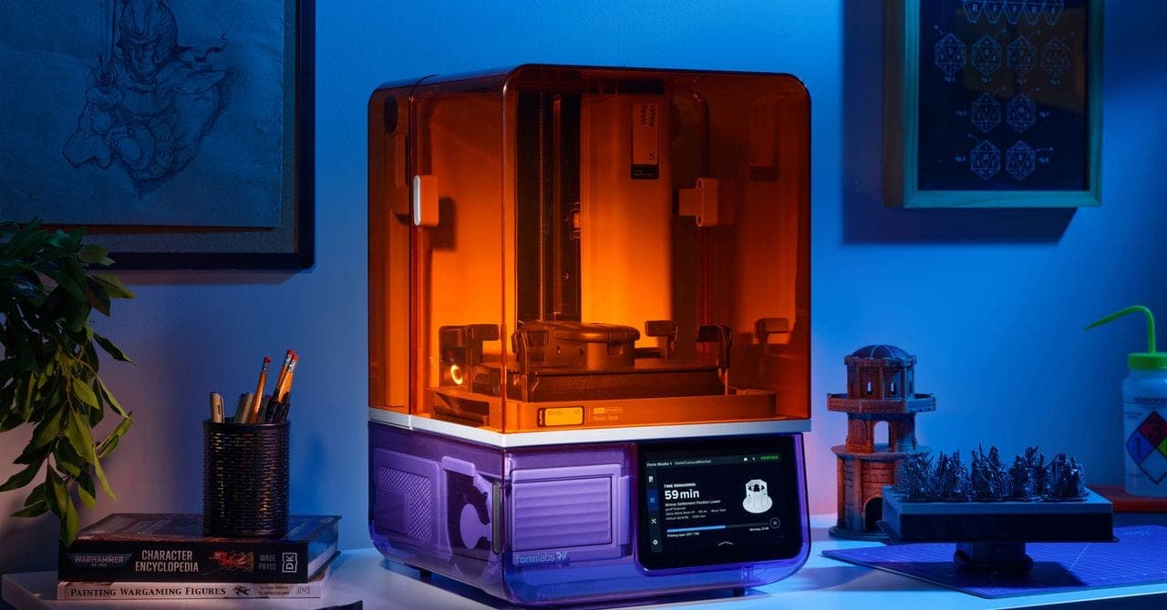A Formlabs Form Studio 1 resin 3D printer with an orange cover and a violet base on a desk, next to printed miniatures, a pencil cup and painting books