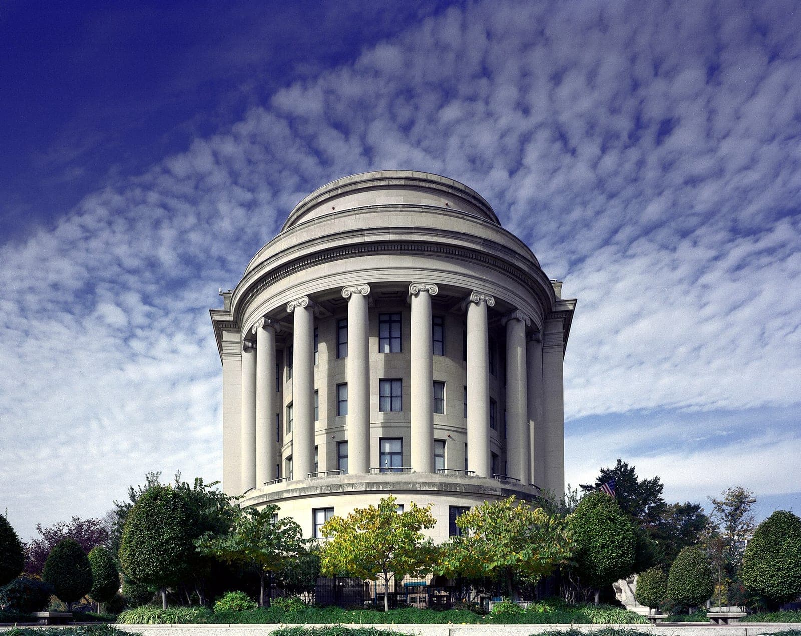 The rounded, columned Federal Trade Commission Building in Washington, D.C., framed by trees under a blue sky with scattered clouds.