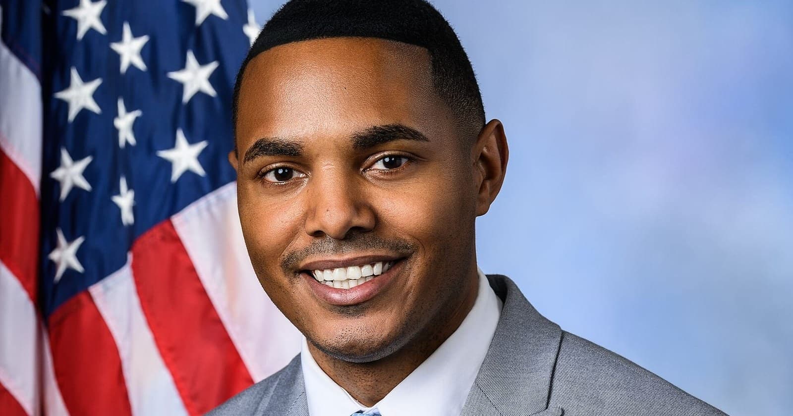 Official portrait of Rep. Ritchie Torres in a gray suit and light blue tie, smiling in front of a US flag