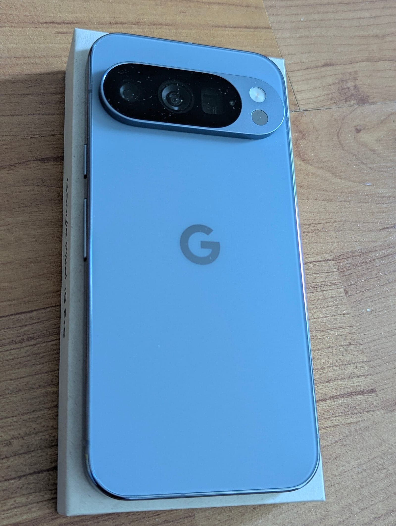 The back of a light blue Google Pixel 10 Pro phone with its camera bar, lying on its box on a wooden table