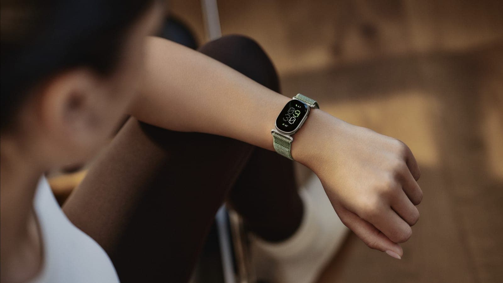 A woman rests her wrist on her knee while wearing the Google Fitbit Edge, a slim capsule-shaped tracker with a green woven band.