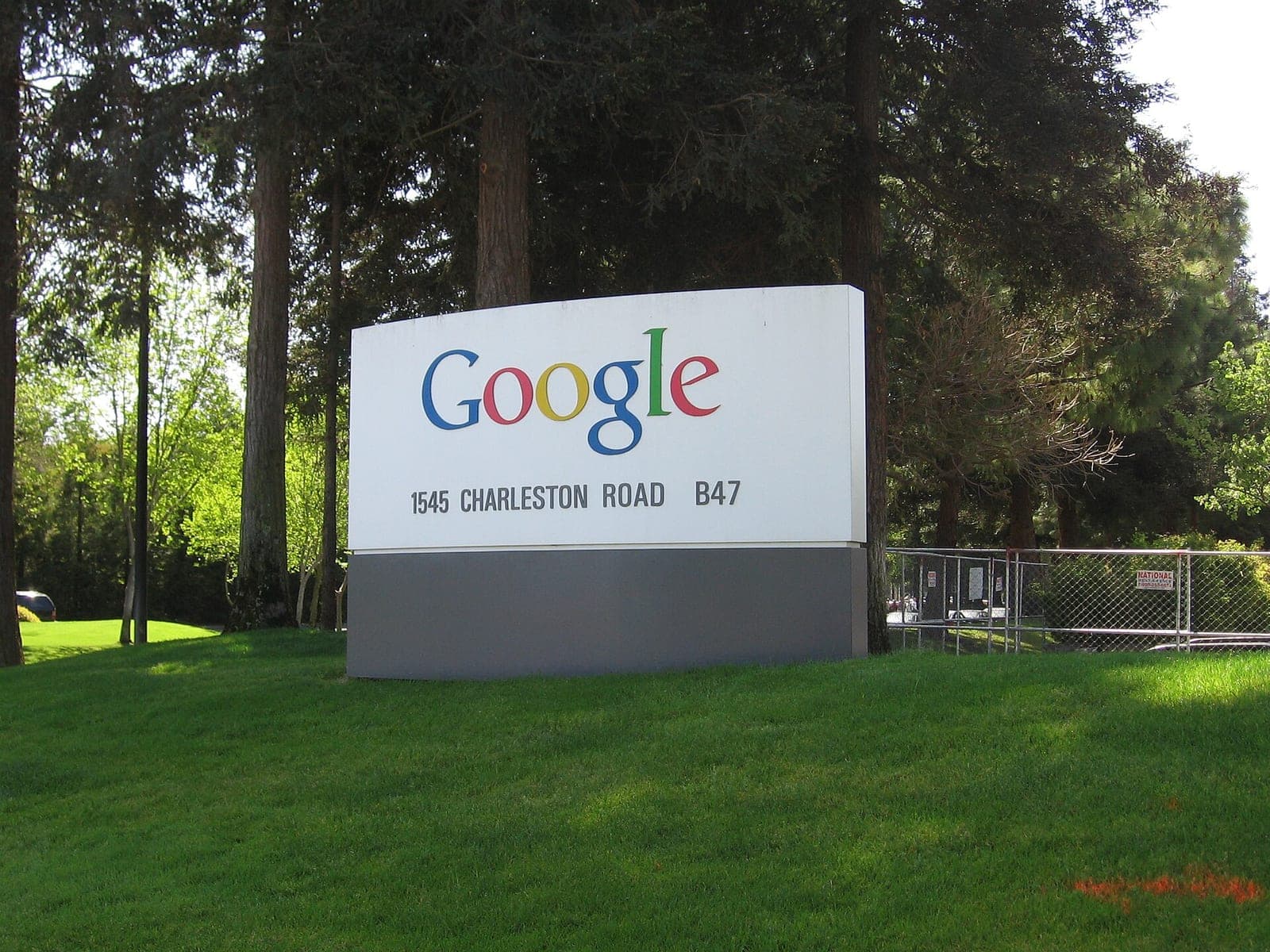A white Google sign with the company's colorful logo and the address 1545 Charleston Road B47, standing on a green lawn in front of tall trees in Mountain View