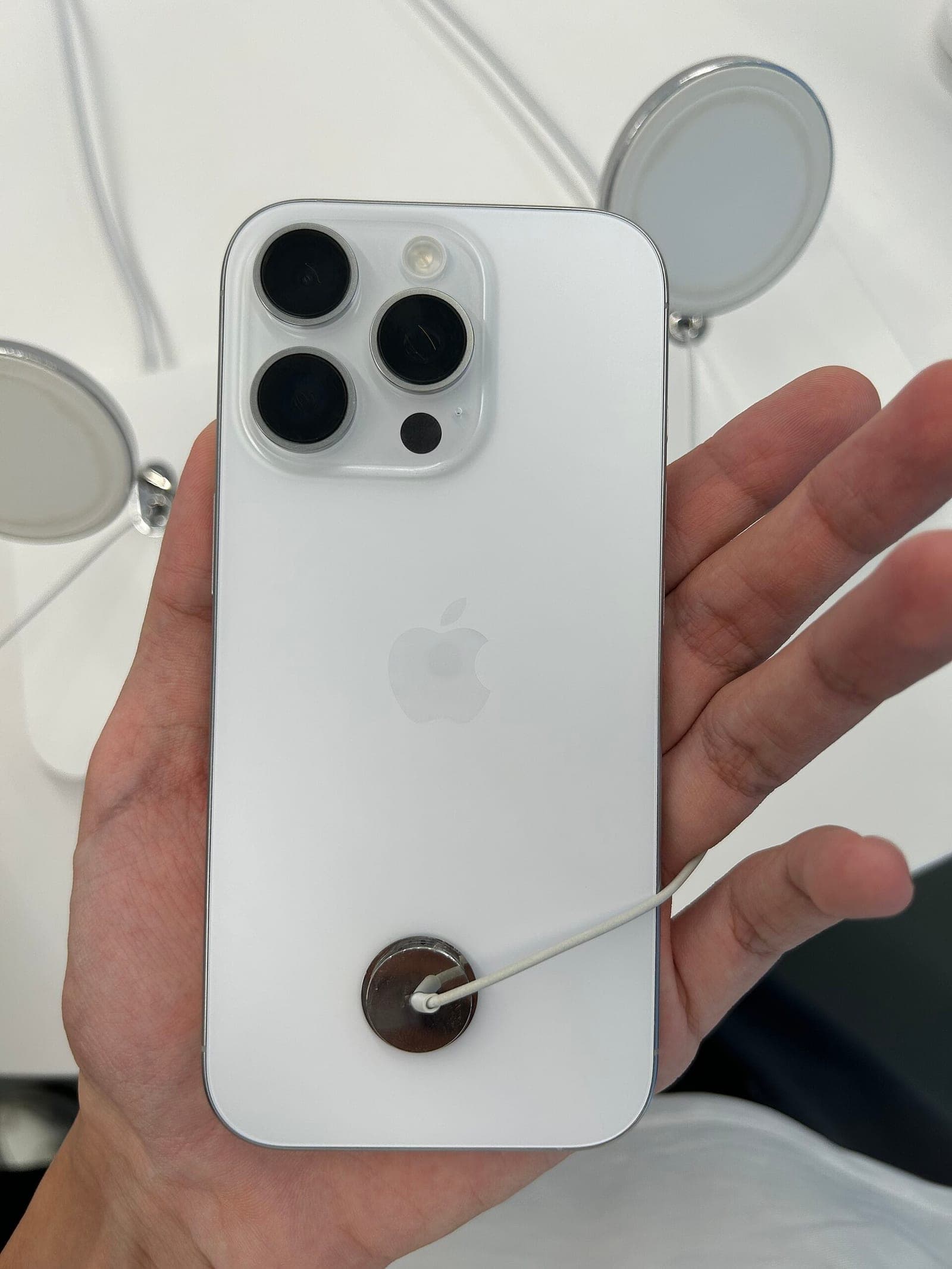 A hand holding the back of a white iPhone 16 Pro with a store security tether attached