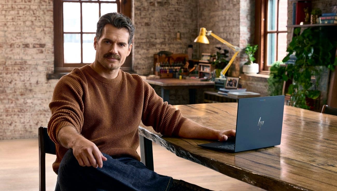 Henry Cavill sits at a wooden table with an HP Googlebook laptop open in front of him