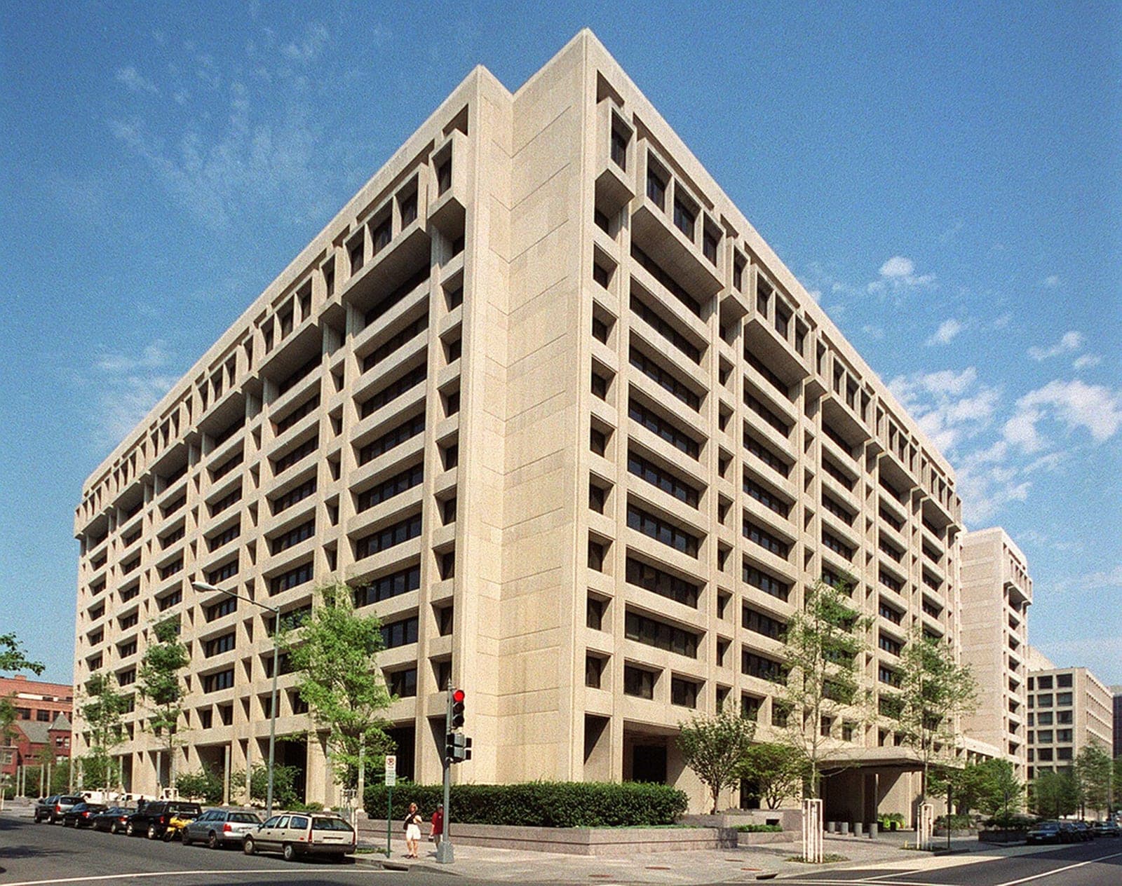 The beige, twelve-story International Monetary Fund headquarters building in Washington, D.C., seen from a street corner under a blue sky, with trees and parked cars in front