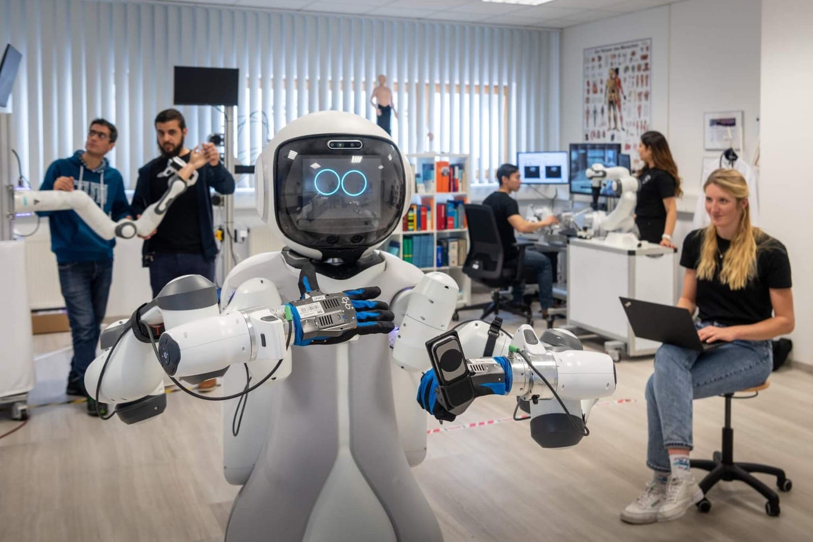 A white humanoid robot with articulated arms standing in a research lab surrounded by researchers