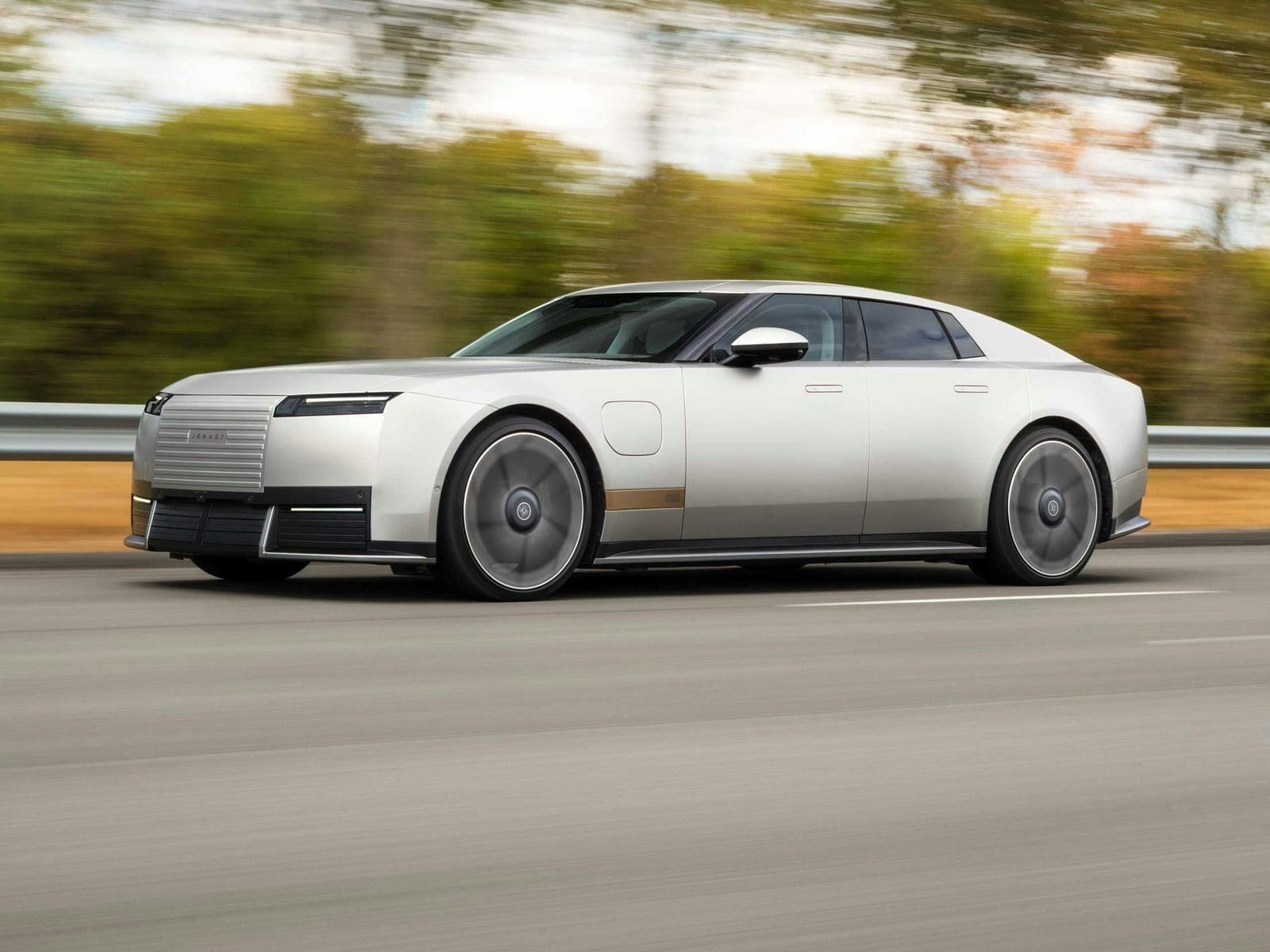 A silver Jaguar Type 01 four-door electric grand tourer driving on a highway with trees blurred behind it