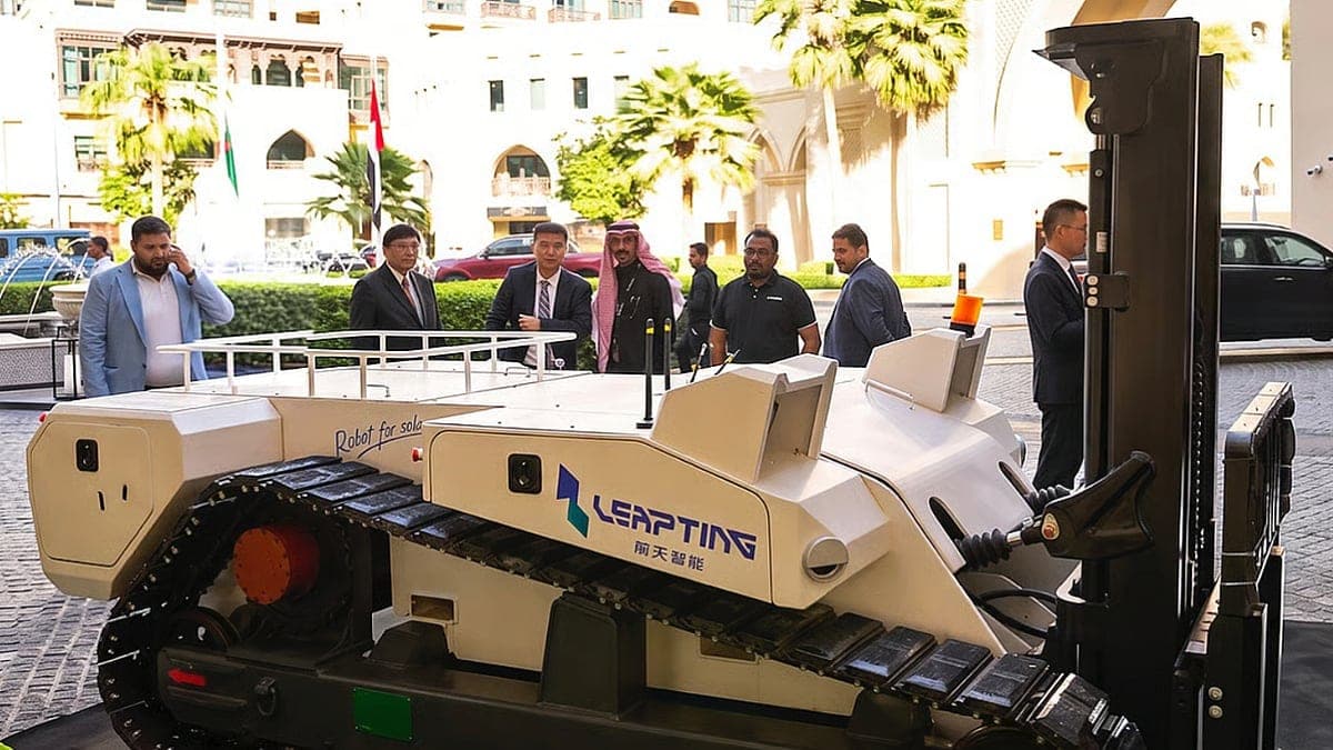 A white LEAPTING tracked forklift robot with a black mast on display outdoors, with a group of people in suits looking at it in front of a building with palm trees