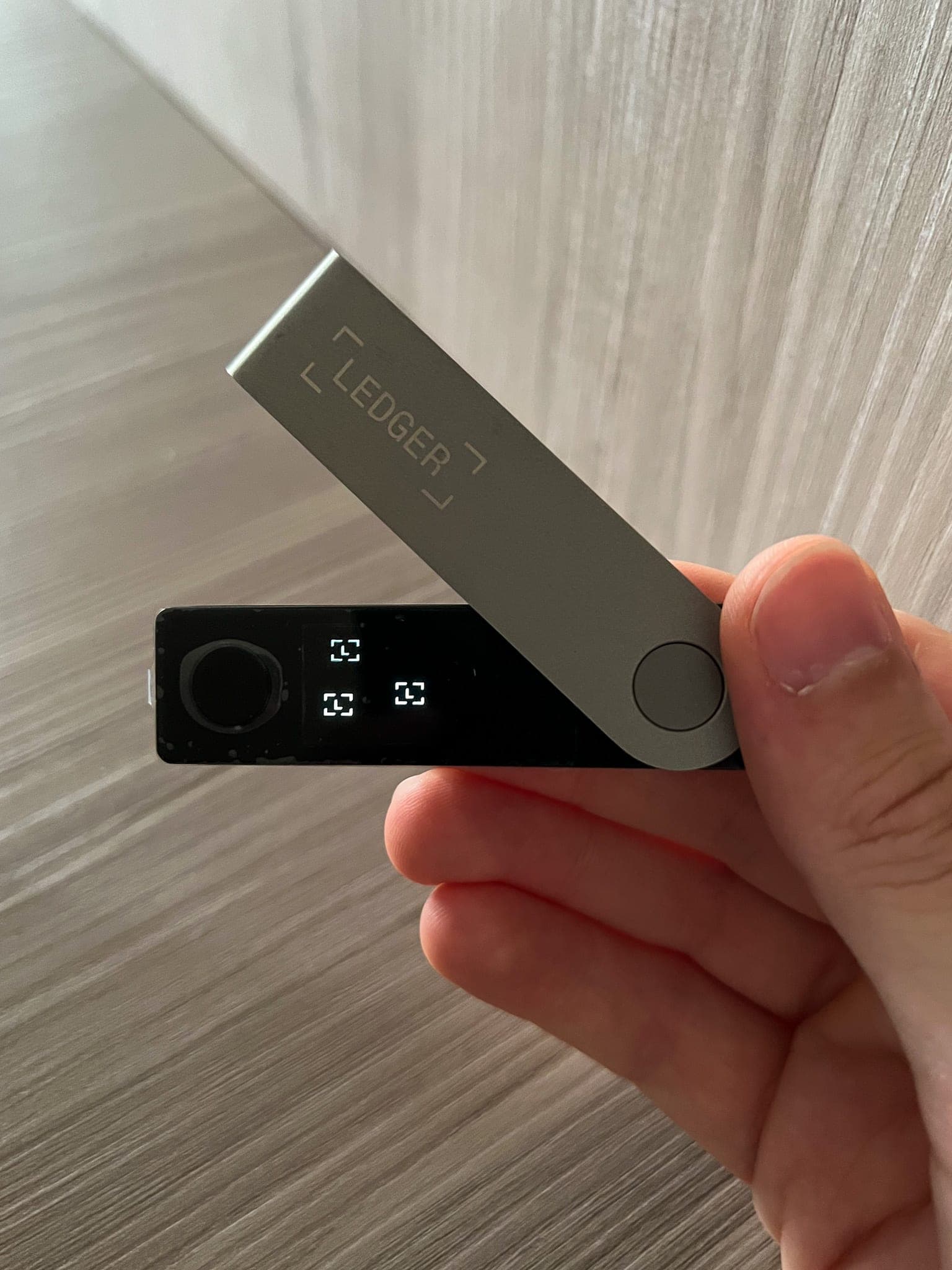 A hand holding an open Ledger hardware wallet, its metal cover swung away from the small black screen
