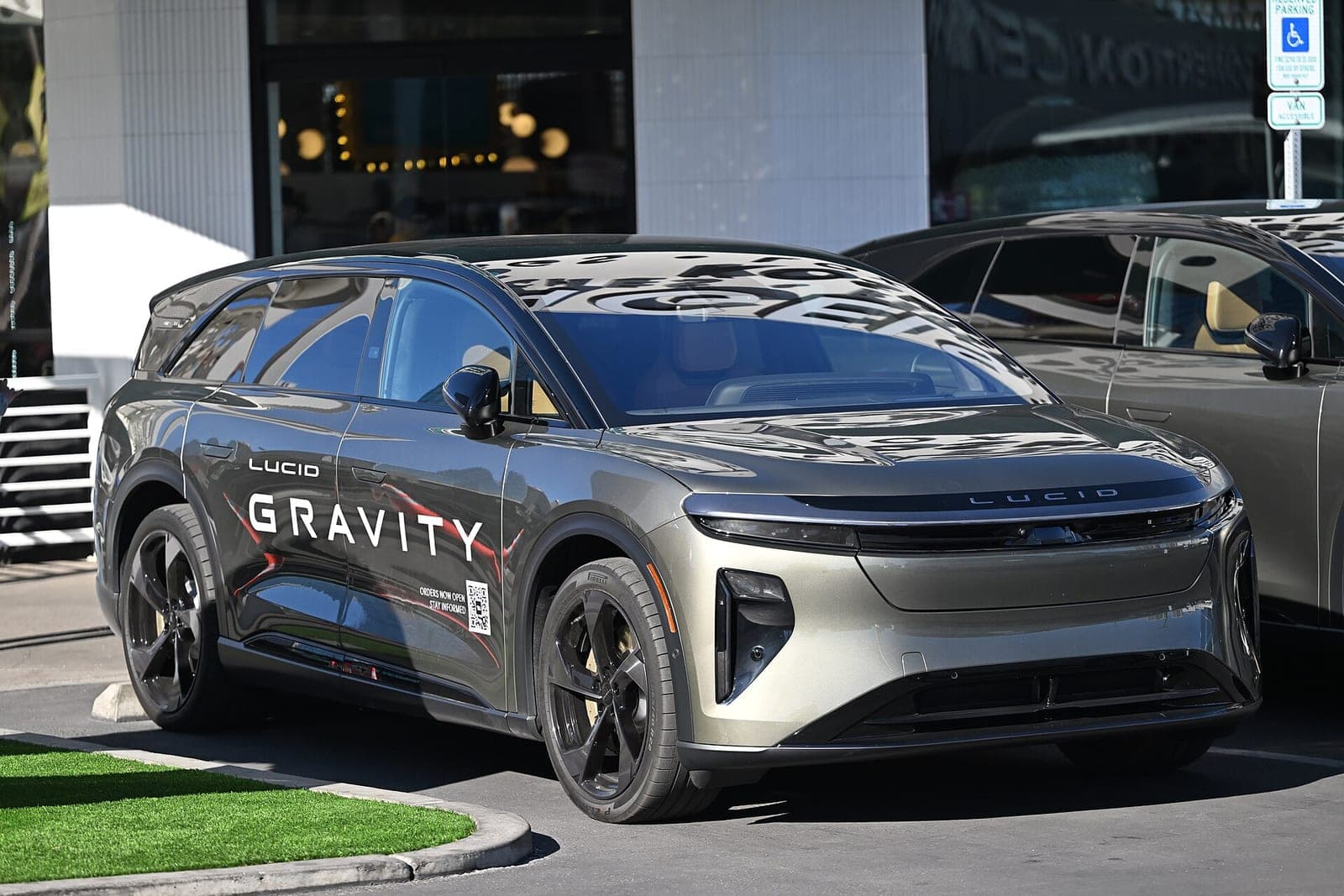 A grey Lucid Gravity electric SUV with 'Lucid Gravity' lettering and an 'orders now open' QR code on its doors, parked outside a glass-fronted building