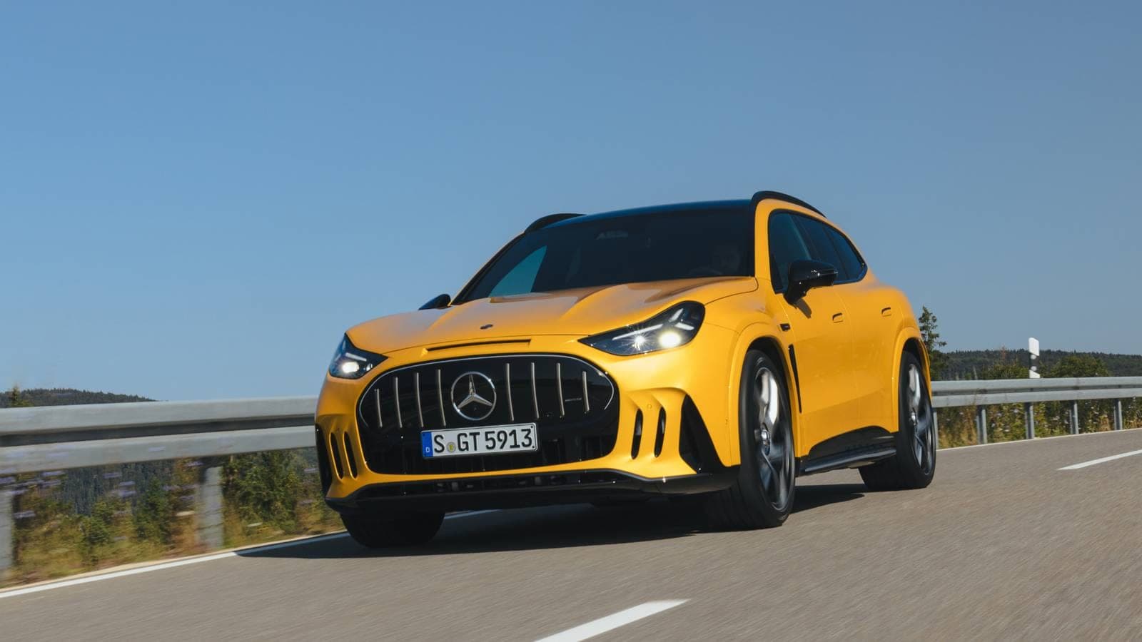 A yellow Mercedes-AMG GT 53 SUV driving on a mountain road with guard rails and a clear blue sky