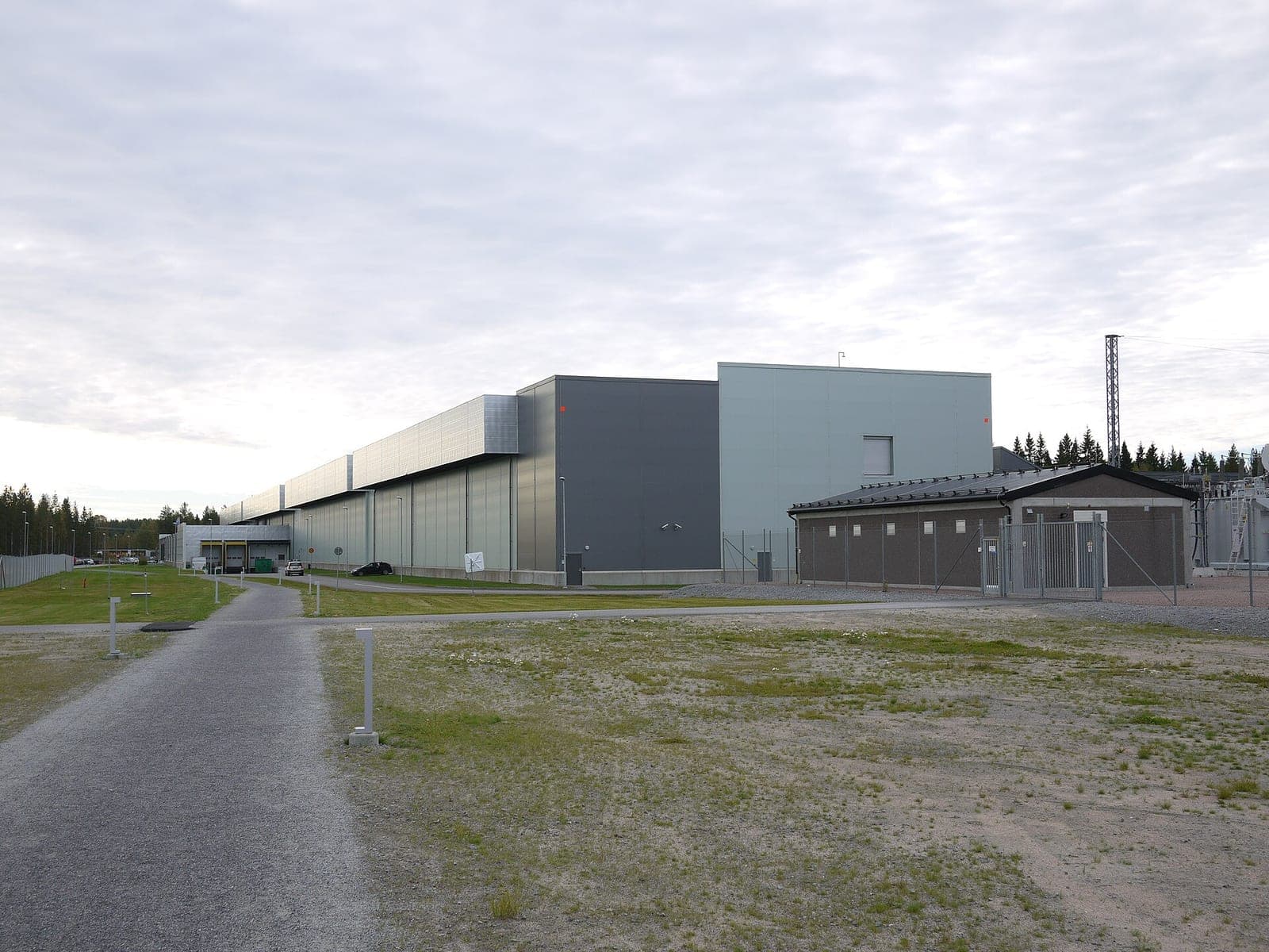 A large industrial data center building with fencing and a gravel lot in front, under an overcast sky
