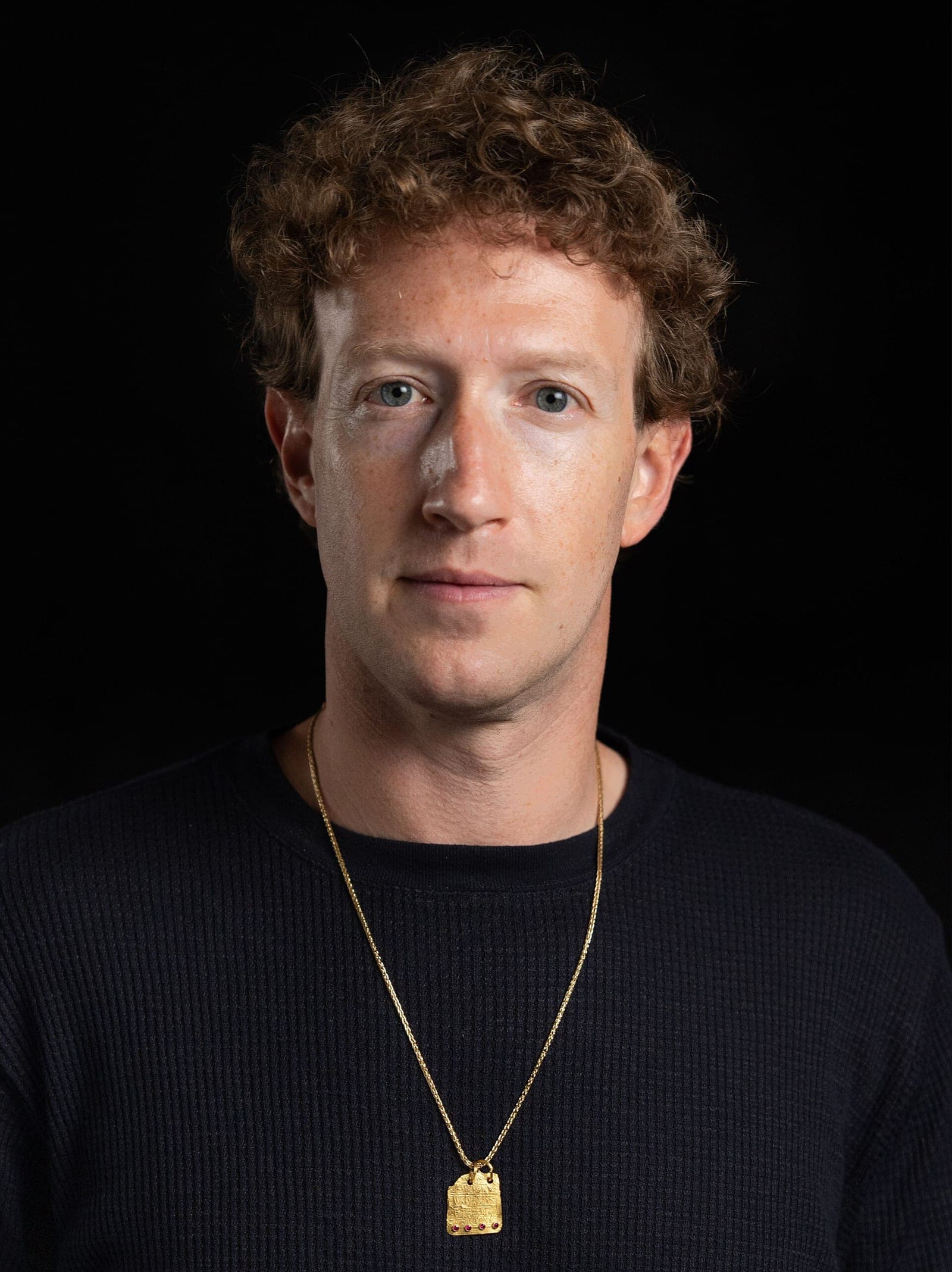 Portrait of Mark Zuckerberg wearing a black sweater and gold necklace against a dark background