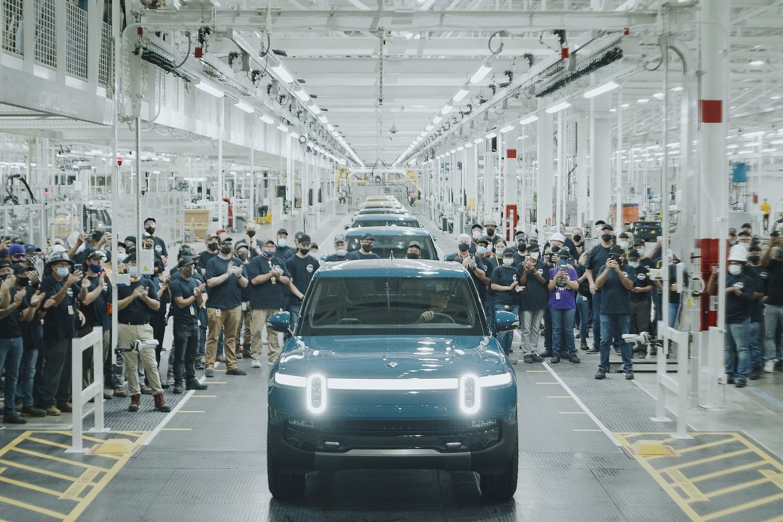 Factory workers in masks applaud along both sides of an assembly line as a blue Rivian R1T pickup with glowing headlights rolls toward the camera at Rivian's plant in Normal, Illinois