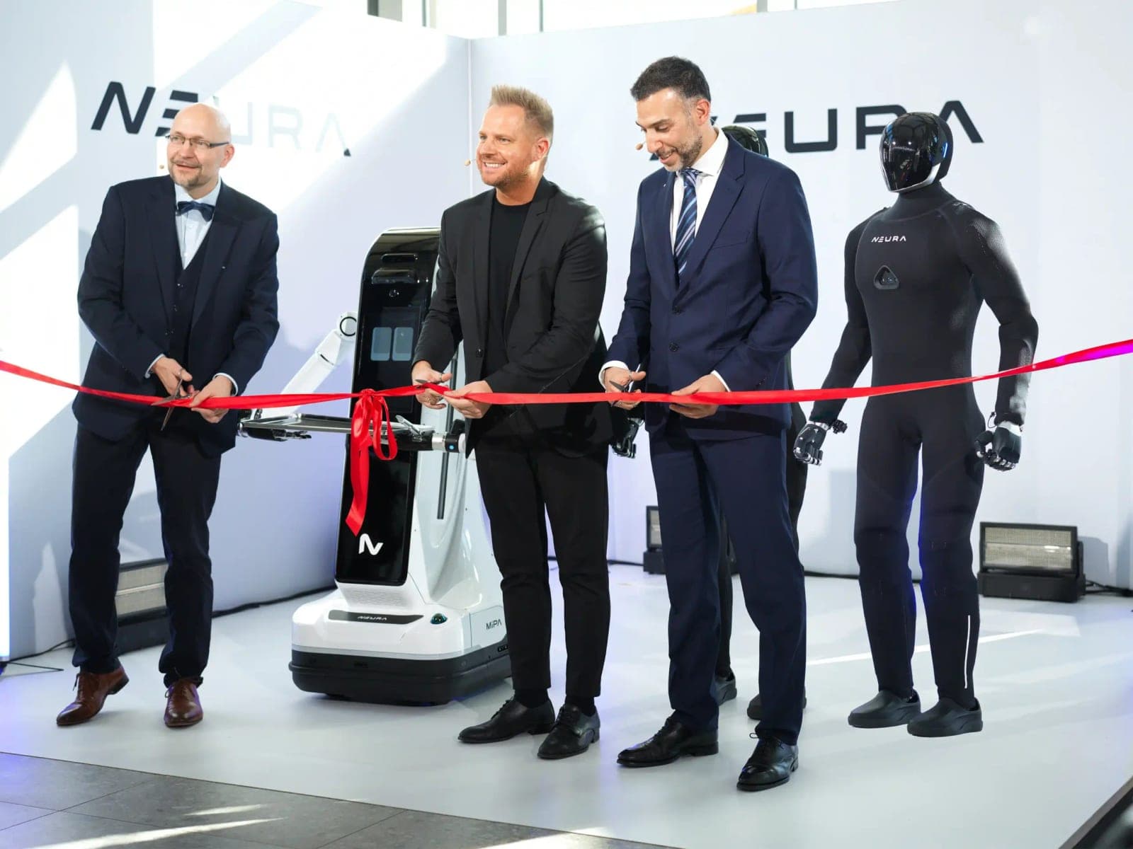 Three men cut a red ribbon at the opening, with a white MiPA mobile robot and a black humanoid in a textile suit standing in front of NEURA logos