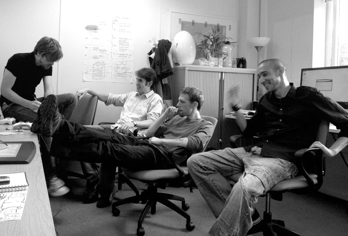 A black and white photo of four Hello Games team members relaxing in office chairs in the studio's early office.