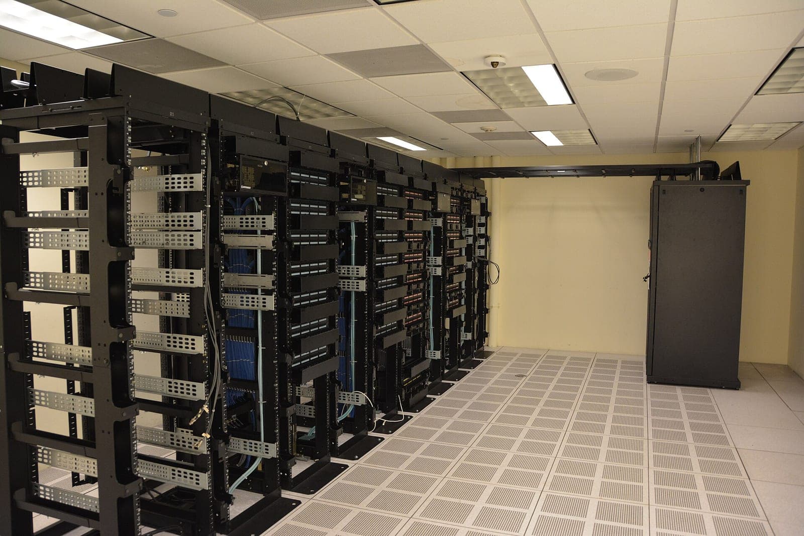 Rows of black server racks with cabling in a data center room with raised floor tiles