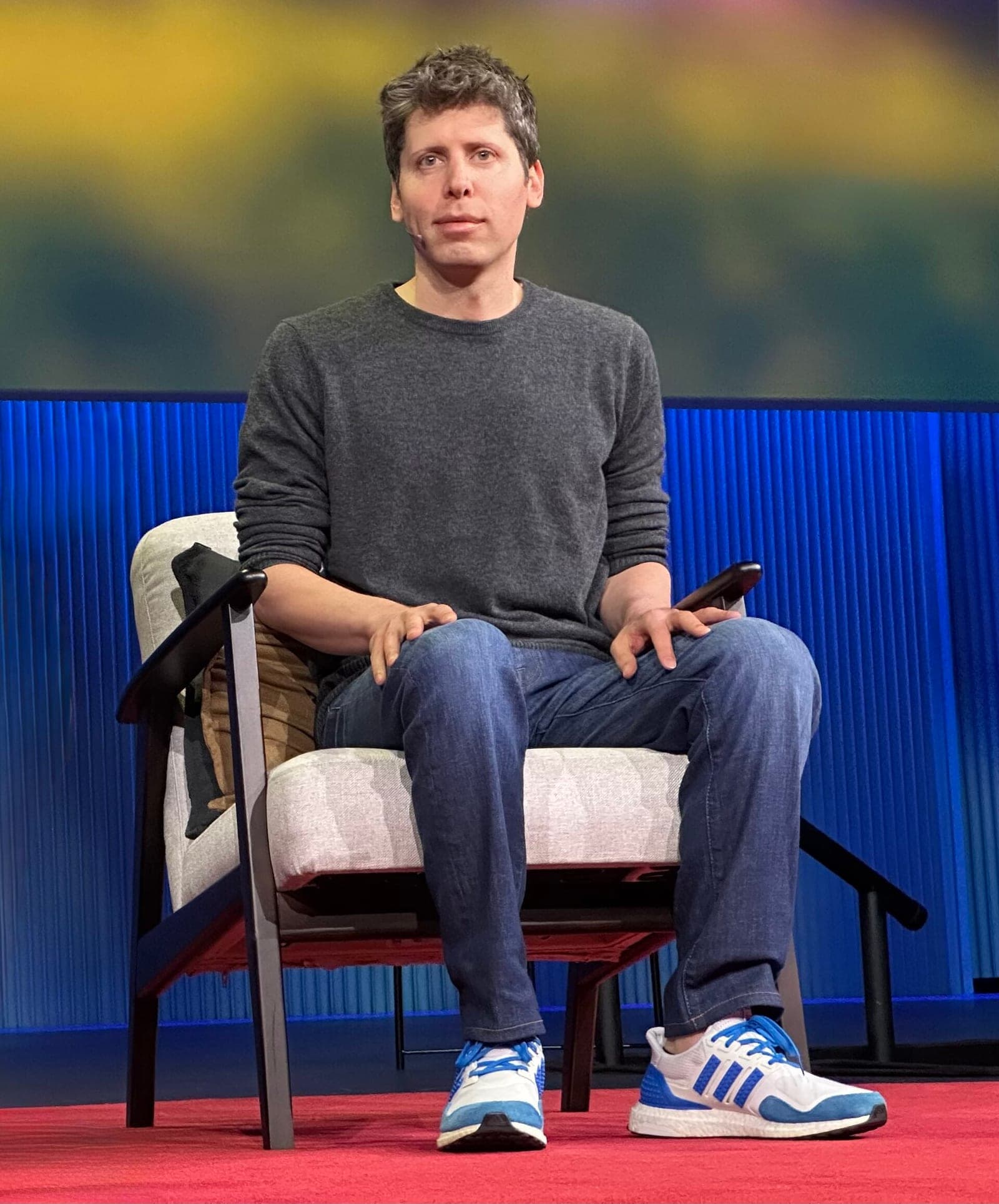 OpenAI CEO Sam Altman seated on stage during a TED talk, wearing a gray sweater and jeans.