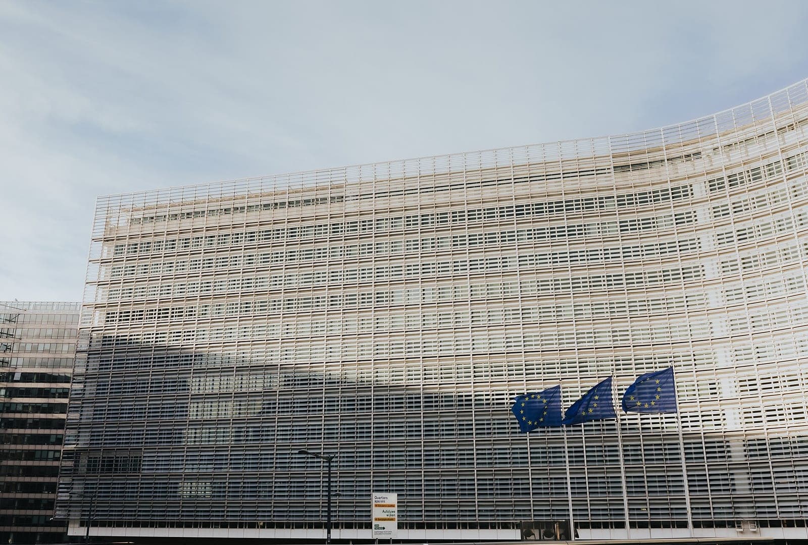 The curved glass facade of the Berlaymont building in Brussels, headquarters of the European Commission, with three EU flags flying in front of it