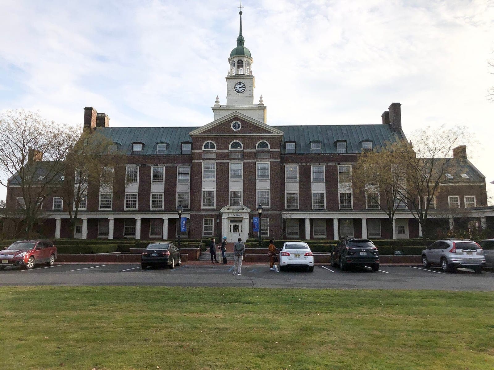 Fuld Hall, a red brick building with a white clock tower, at the Institute for Advanced Study in Princeton, New Jersey, with cars parked in front