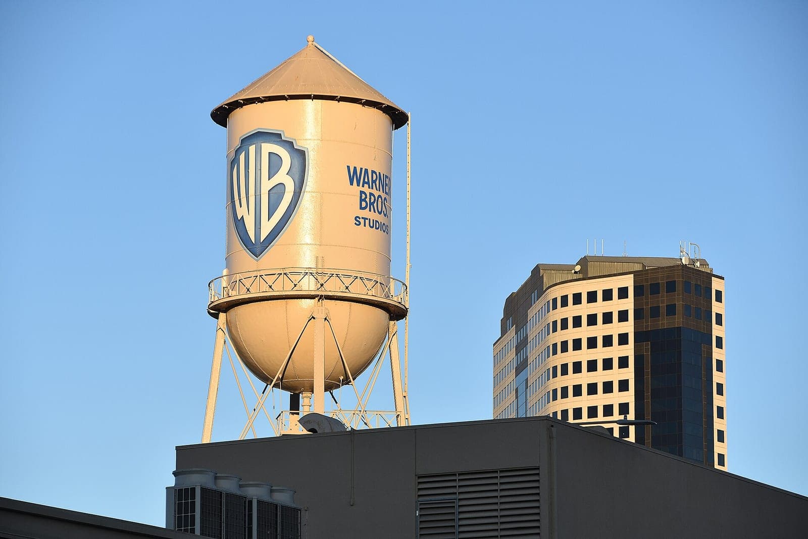 The Warner Bros. Studios water tower with the WB shield logo against a clear blue sky, next to an office tower