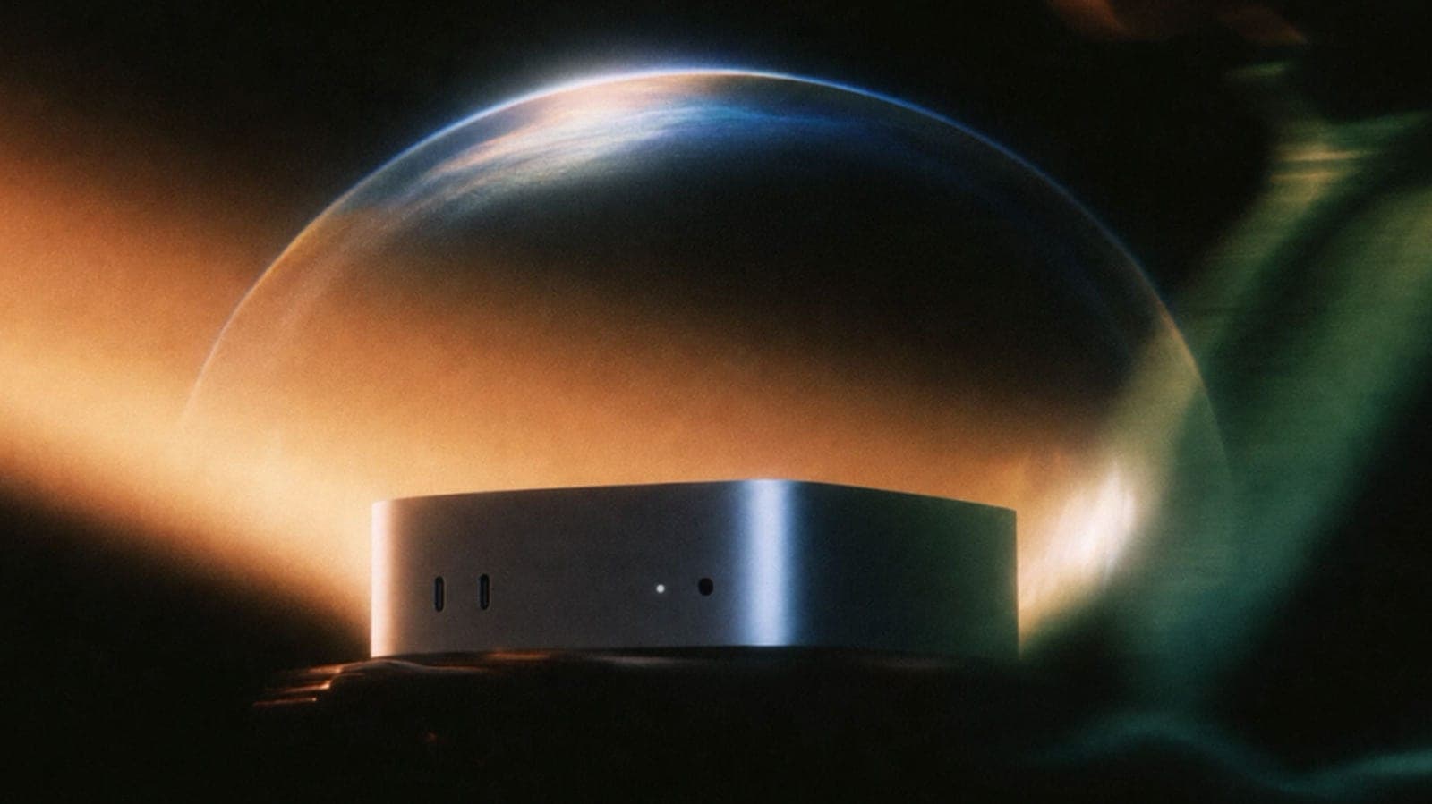 A silver Mac mini sits on a dark surface under a clear glass dome, lit by warm orange light against a dark green and black background