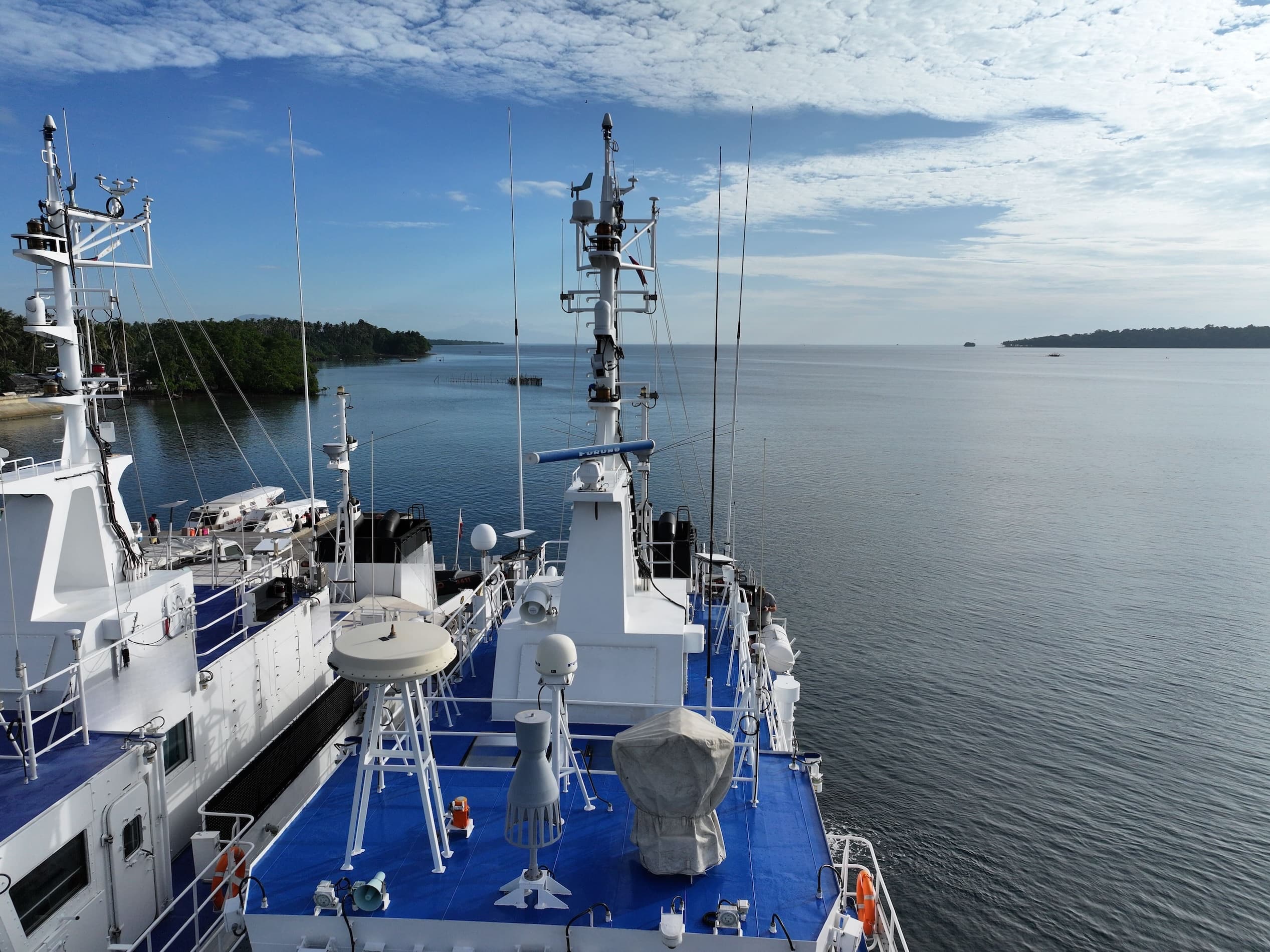 The mast of a ship equipped with multiple sensor and radio antennas, docked near a tropical coastline.