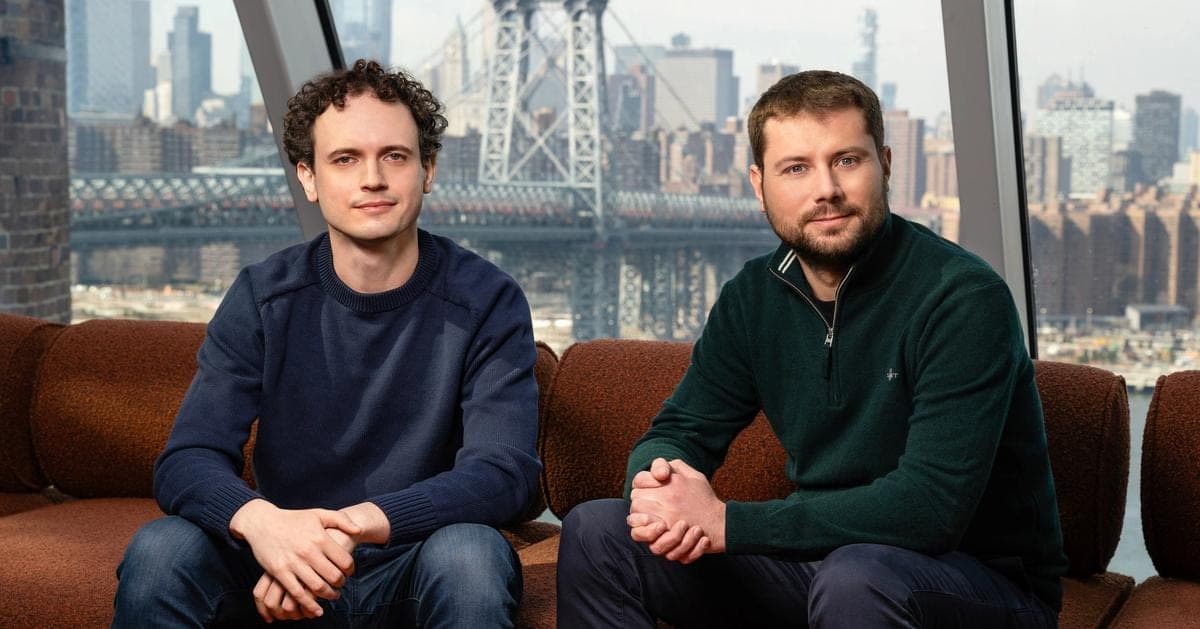 Reflection AI co-founders Misha Laskin and Ioannis Antonoglou sitting on a rust-colored couch in front of a window overlooking the Williamsburg Bridge and the New York skyline