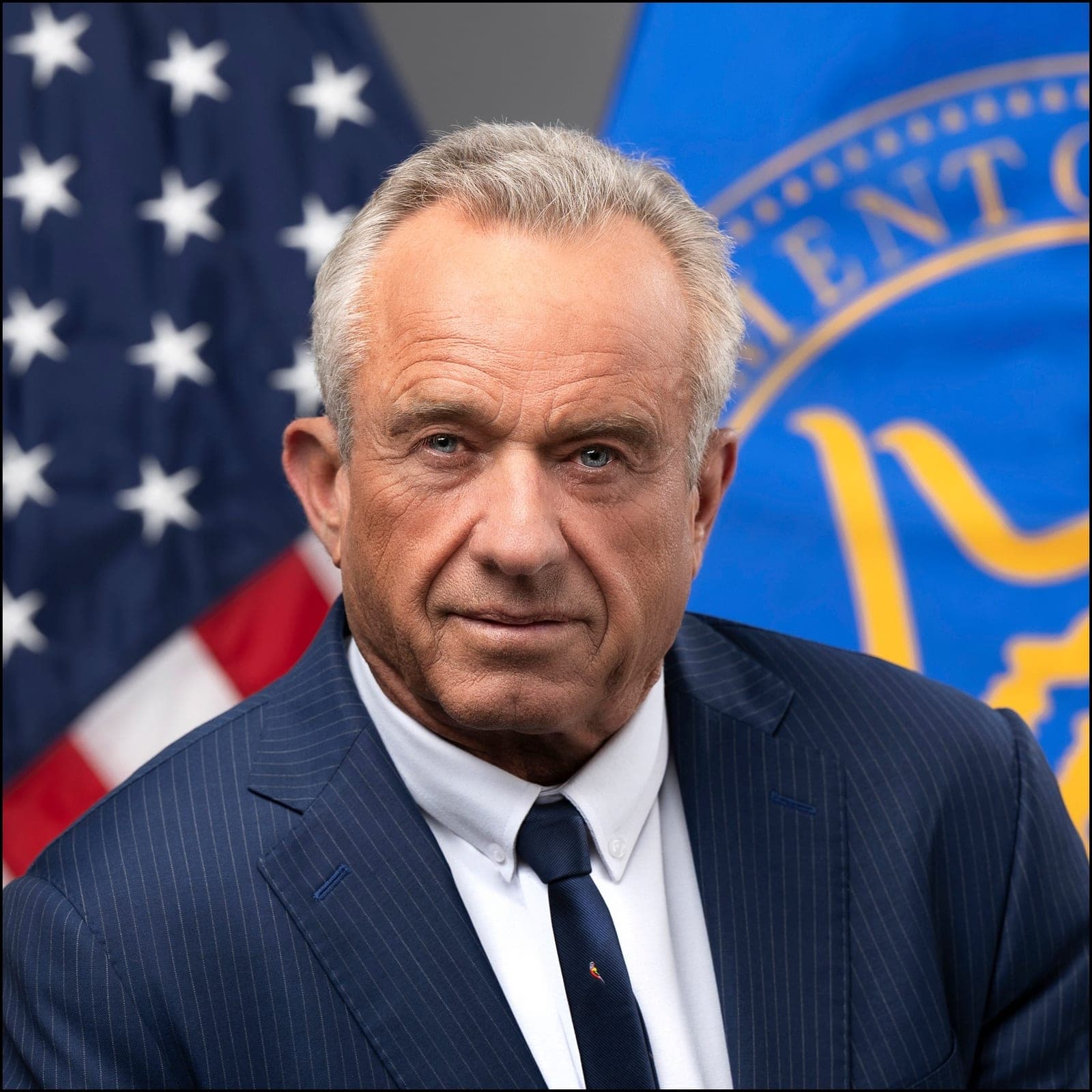 Official portrait of Robert F. Kennedy Jr. in a suit and tie, with American and Department of Health and Human Services flags behind him