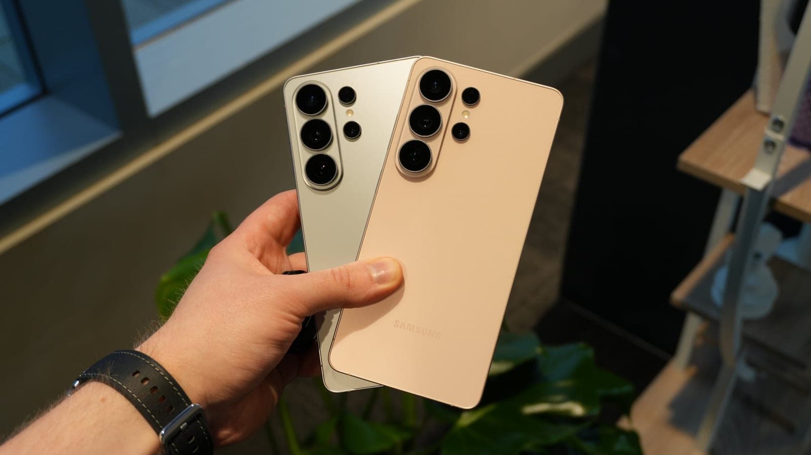 A hand holding two Galaxy S26 Ultra phones in silver and rose gold colors, showing their camera arrays
