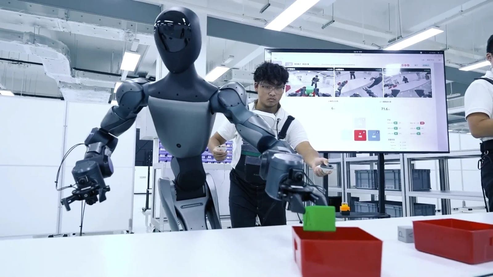 A dark gray humanoid robot reaches toward a green block above a red bin while a technician in a work vest guides it with handheld controllers, with a monitor of camera views behind them