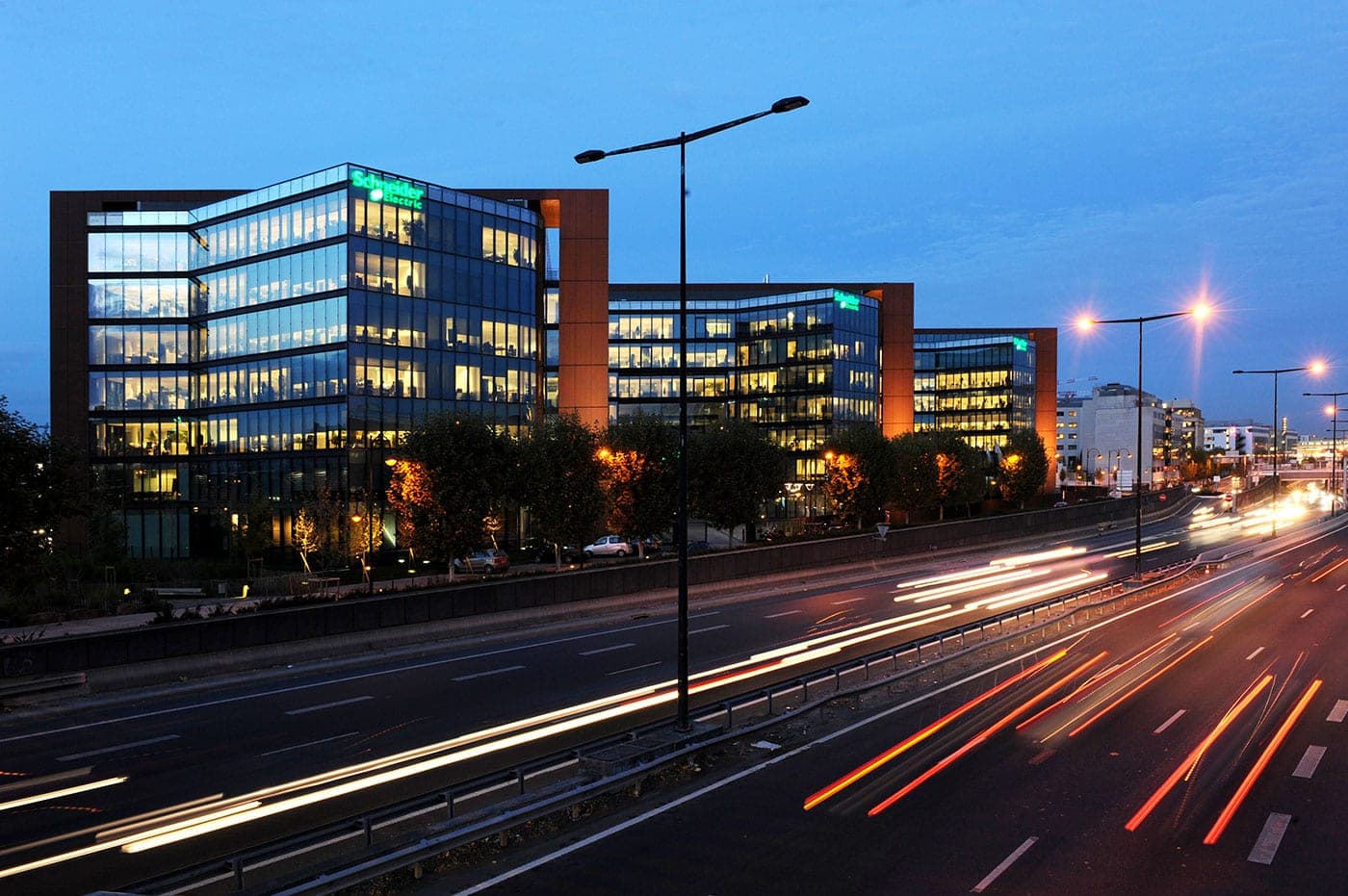 Schneider Electric's glass-fronted headquarters in Rueil-Malmaison, France, lit up at dusk with green Schneider Electric signs, beside a highway with car light trails