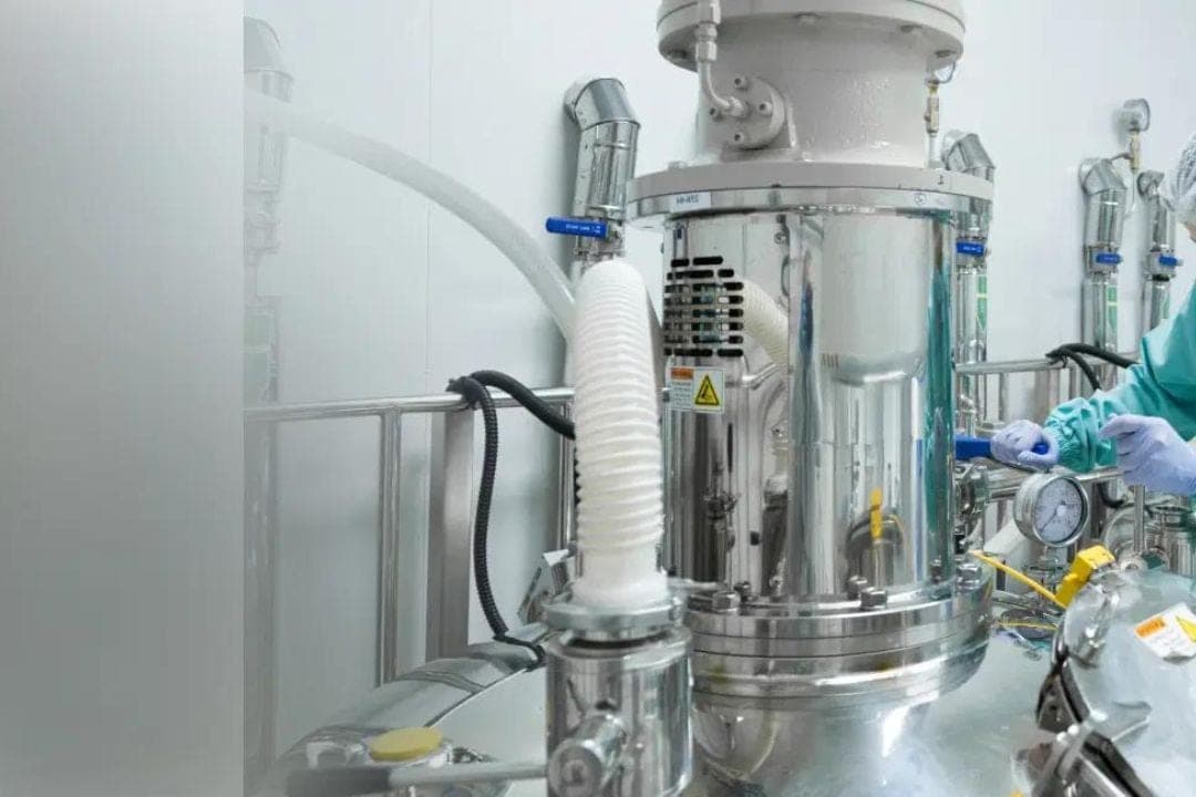 A stainless steel chemical reactor with white flexible hoses and gauges in a clean production room, with a worker in a green gown and blue gloves adjusting a valve on the right