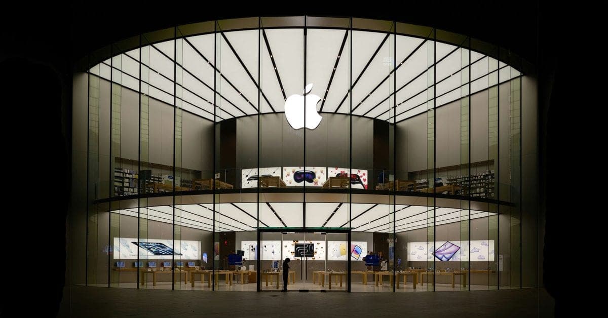 A glass-fronted Apple Store at night, lit from inside, with tables of devices on two floors