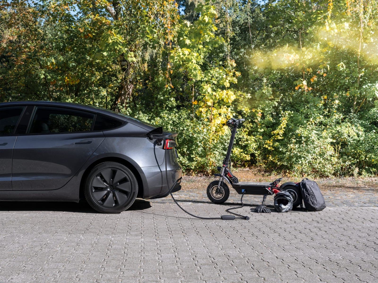 A gray Tesla Model 3 parked on paving stones with a cable running from its charge port to an electric scooter, a helmet and a backpack beside it.