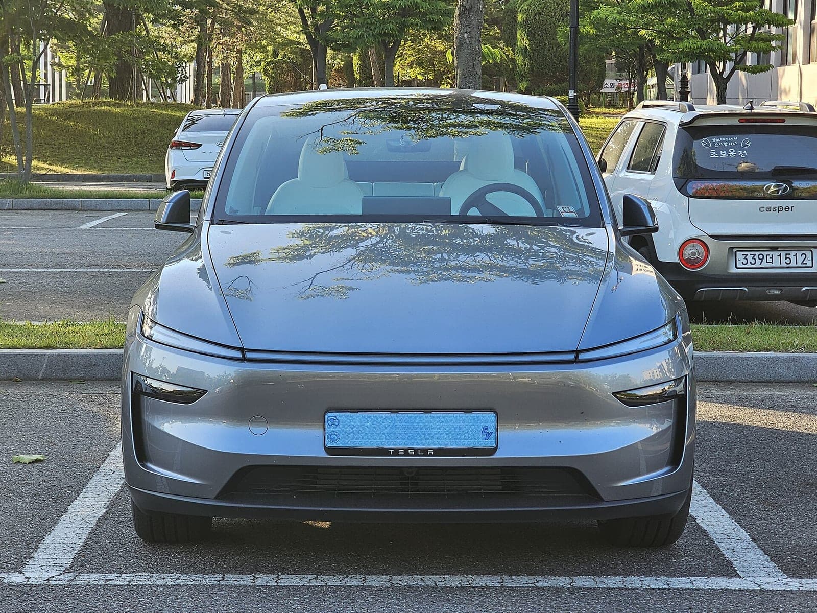 Front view of a silver Tesla Model Y parked in a parking lot with trees behind it