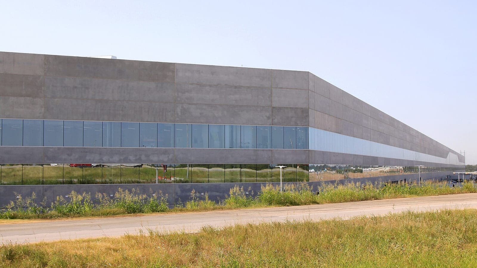 Long concrete and glass wall of Tesla's Gigafactory Texas building seen from a road, with tall grass in the foreground