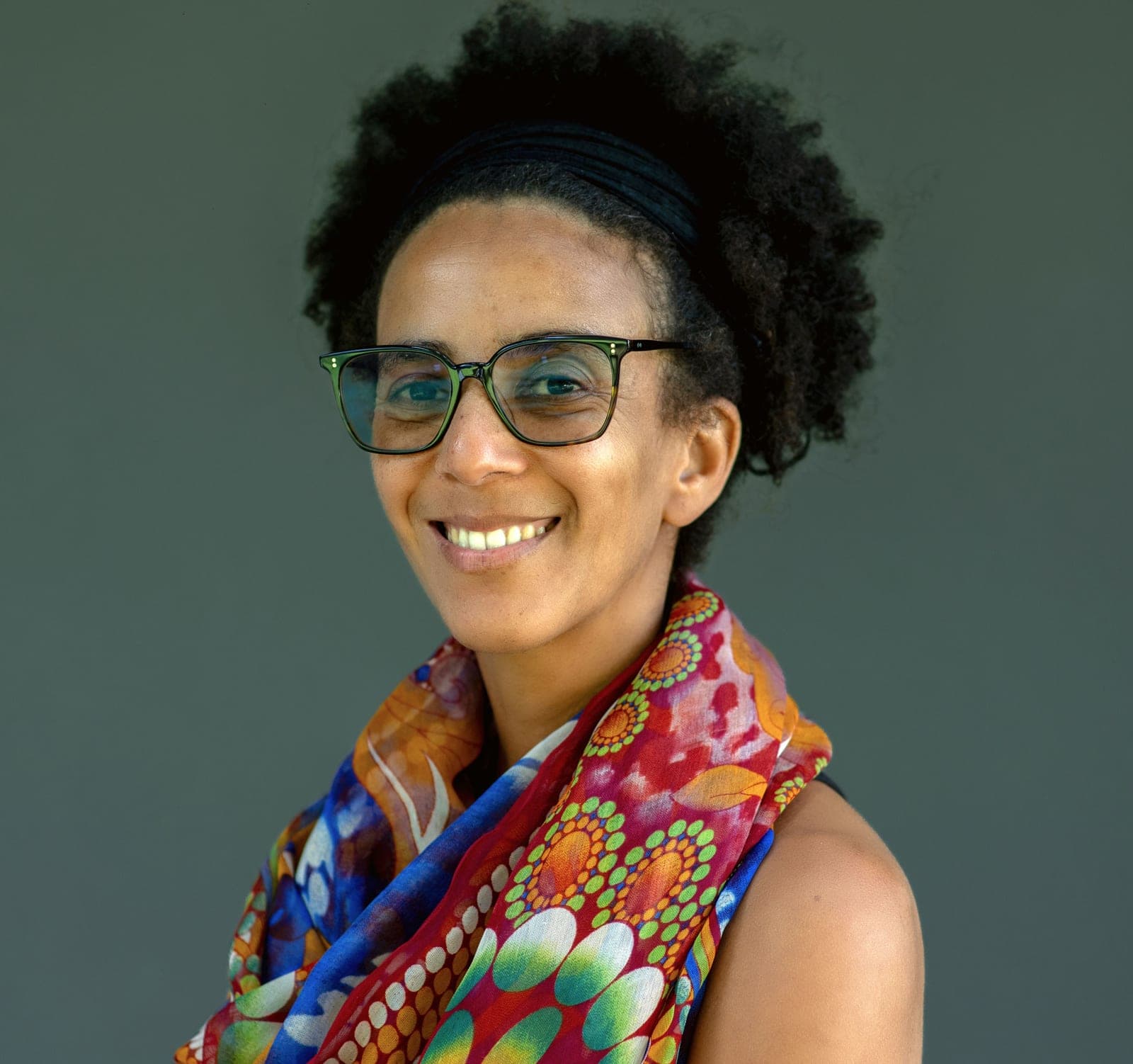 Portrait of AI researcher Timnit Gebru, wearing glasses and a colorful scarf, smiling against a plain gray background.