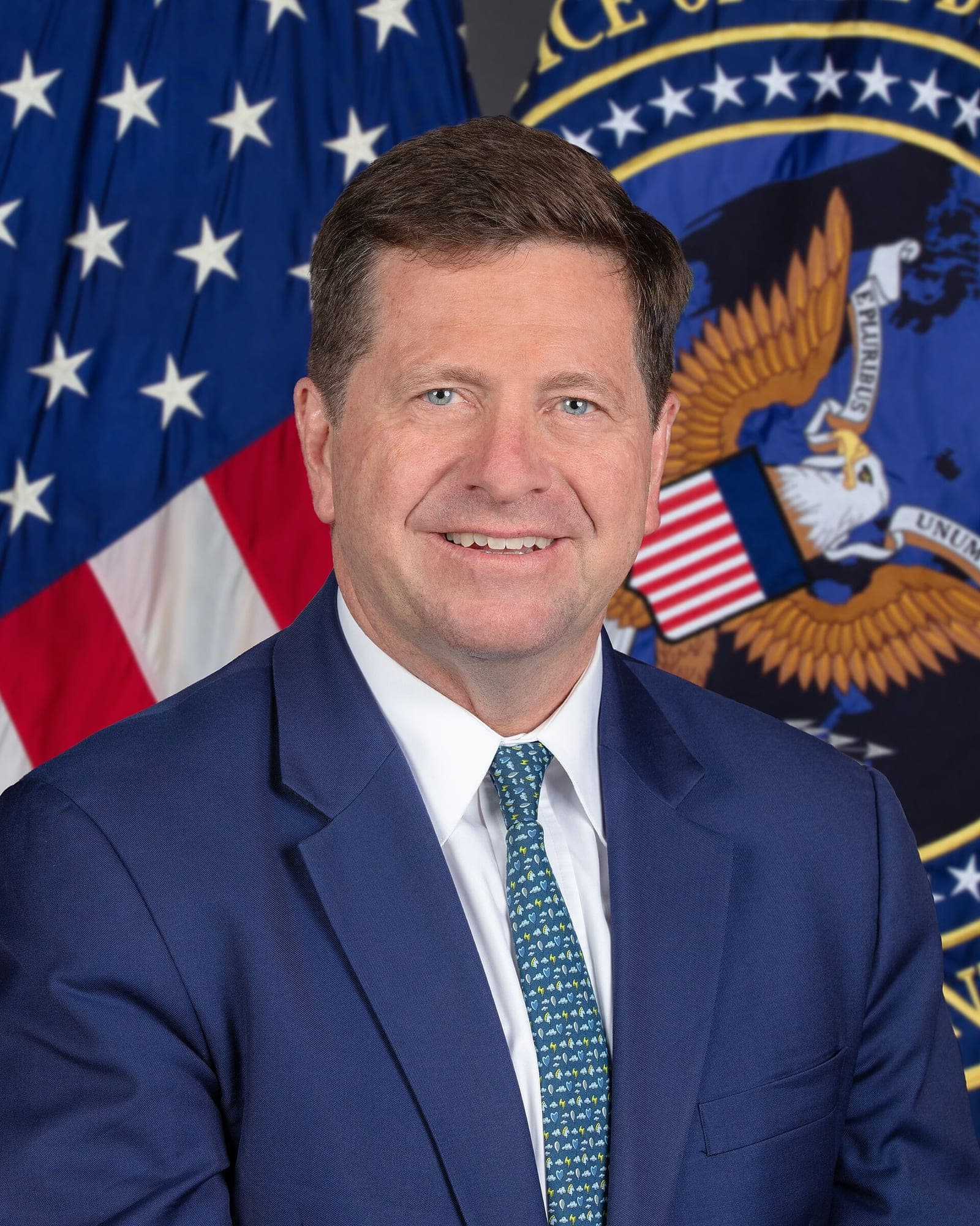 Official portrait of Director of National Intelligence Jay Clayton, smiling in a navy suit and patterned blue tie in front of the US flag and the seal of the Office of the Director of National Intelligence