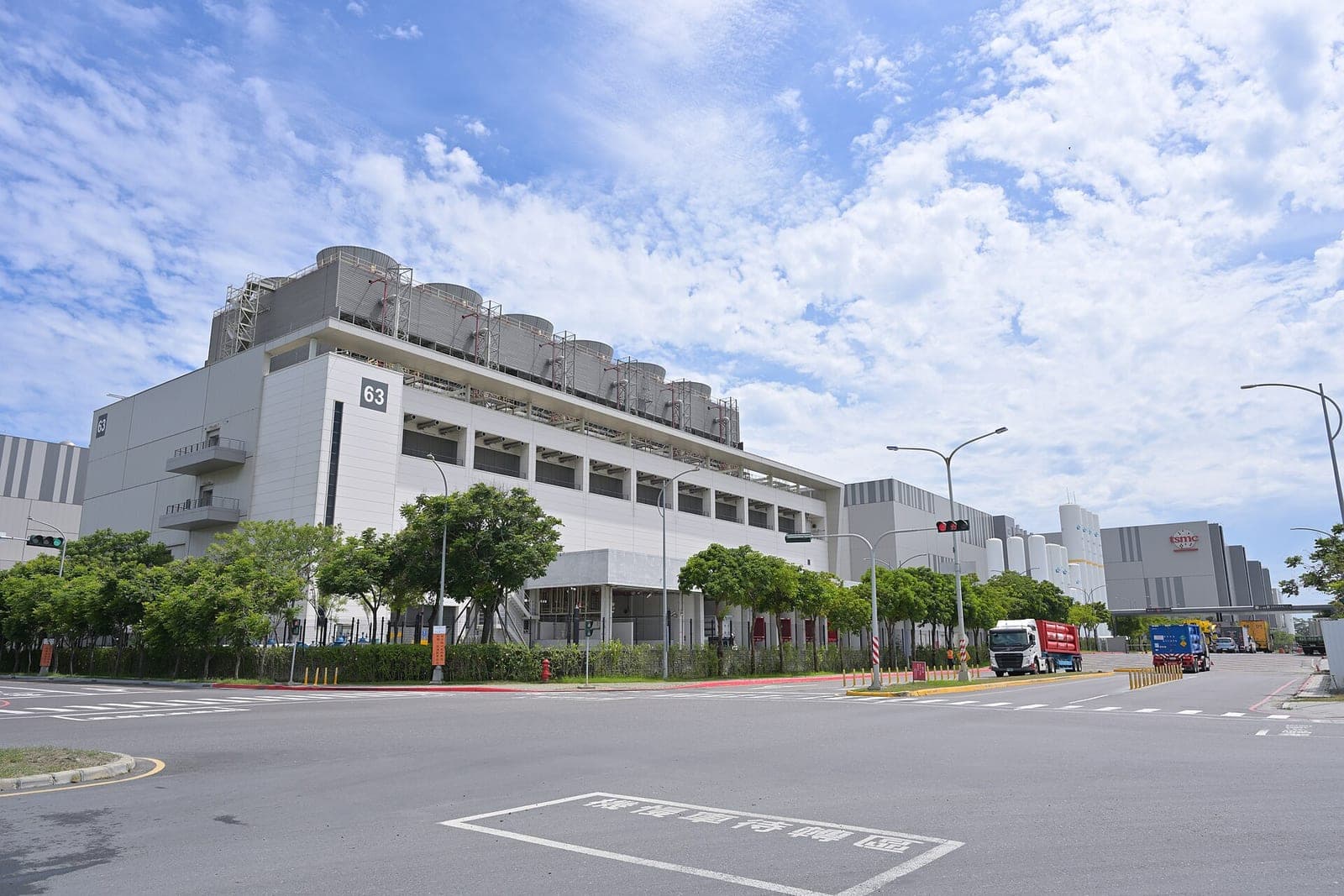 A large white TSMC factory building marked 63 with rows of cooling towers on its roof, trees along the street in front and trucks driving past under a blue sky with white clouds
