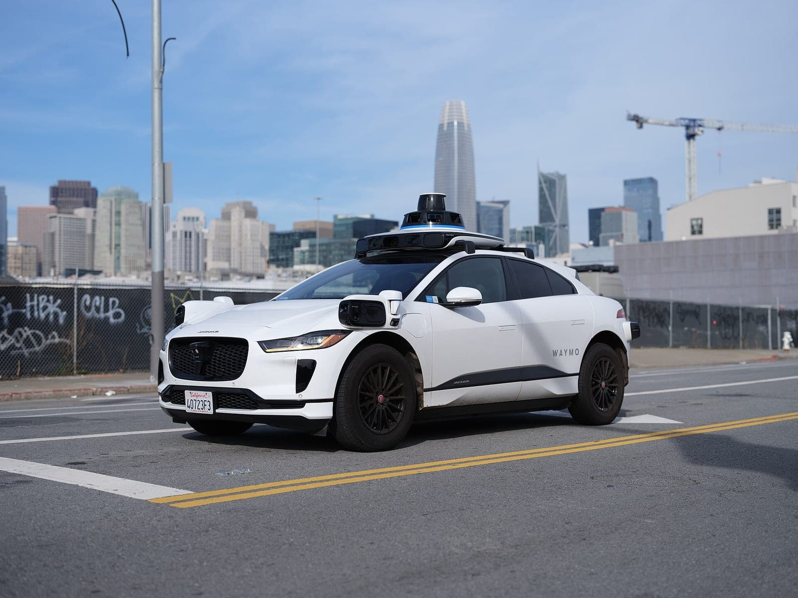 A white Waymo Jaguar I-Pace robotaxi with a sensor dome on its roof parked on a street, with the San Francisco skyline behind it