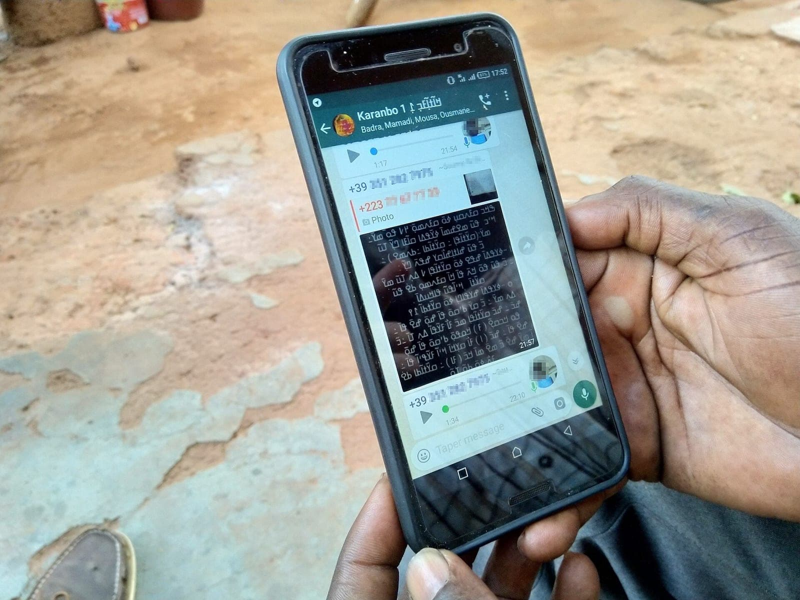 A man holds a smartphone showing a WhatsApp group conversation, with phone numbers blurred
