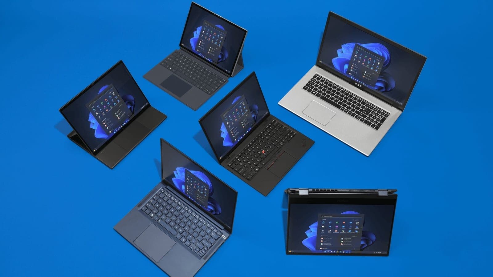 Six Windows 11 laptops and tablets of different sizes and colors arranged on a bright blue surface, each showing the Start menu