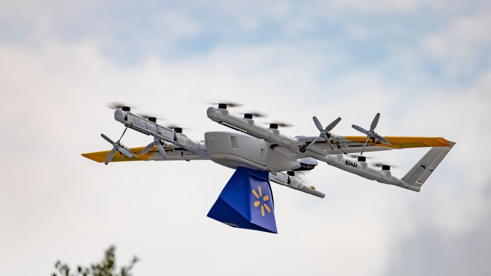 A white Wing delivery drone with yellow wing tips and multiple propellers flies against a cloudy sky, carrying a blue Walmart-branded package beneath it